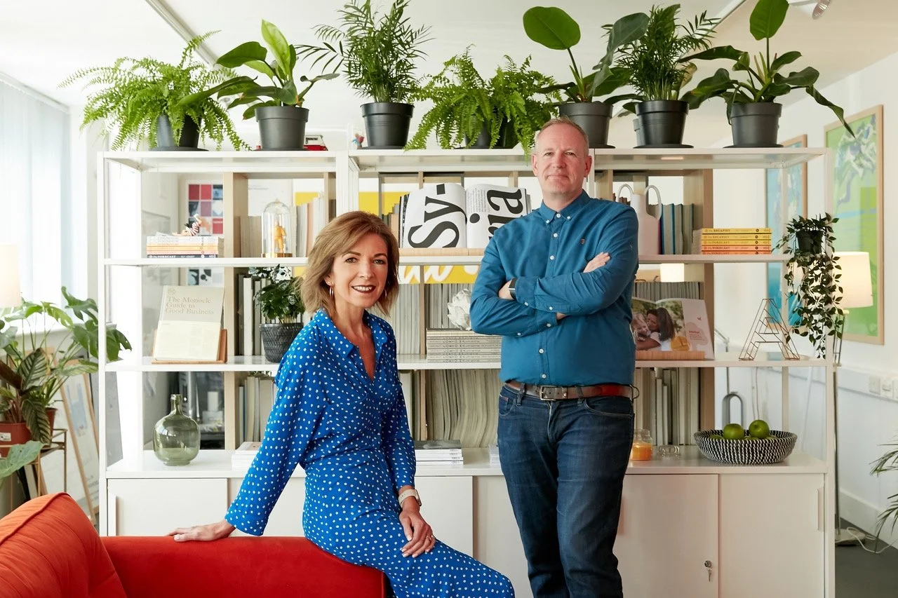 A woman in a blue polka dot dress and a man in a blue shirt with dark jeans pose in a well-lit, cozy living room with a modern aesthetic. They are in front of a white shelf filled with books, decorative items, and potted plants, with large leafy plants on top of the shelf. The woman is sitting on a red sofa and smiling, while the man stands with his arms crossed, smiling at the camera.