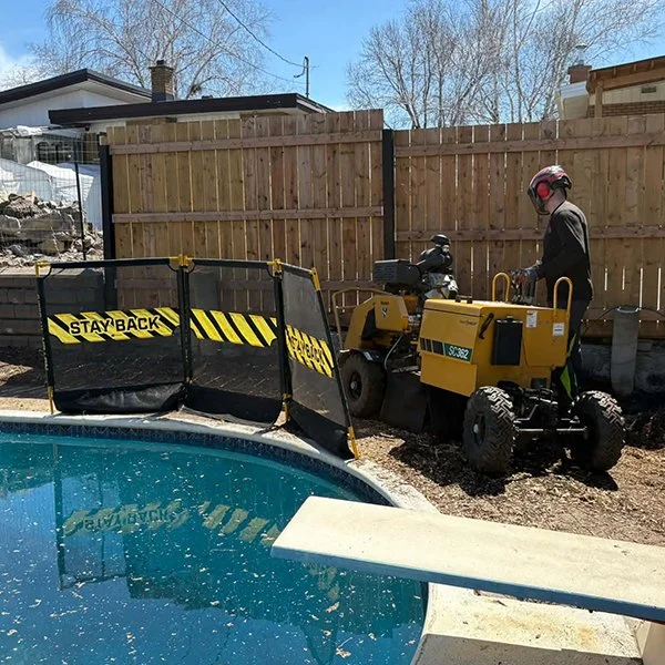 Chauffeur de machine de construction portant un casque de sécurité, utilisant une boue tondeuse près de la piscine, zone clôturée avec panneau 'STAY BACK'.