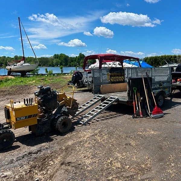 Matériel de nettoyage et de travaux de maintenance près d'un plan d'eau avec voiliers, outils, une remorque et un moteur de bateau.