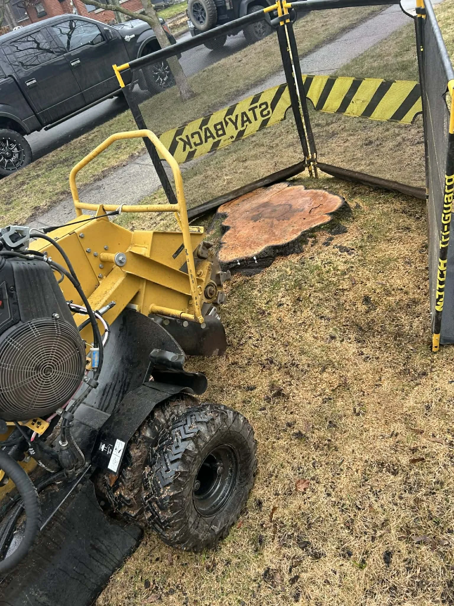 Une machine de coupe d'arbres en train de couper un tronc d'arbre, avec une barrière de sécurité de couleur jaune et noire. La scène se déroule en plein air, sur une pelouse avec des voitures garées en arrière-plan.