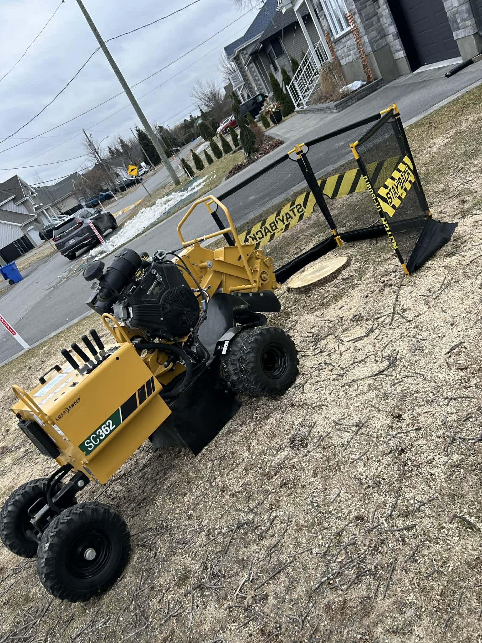 Une machine de déneigement stationnée sur la pelouse d'un quartier résidentiel, avec des maisons en arrière-plan, de la neige en tas sur le trottoir, et un ciel nuageux.