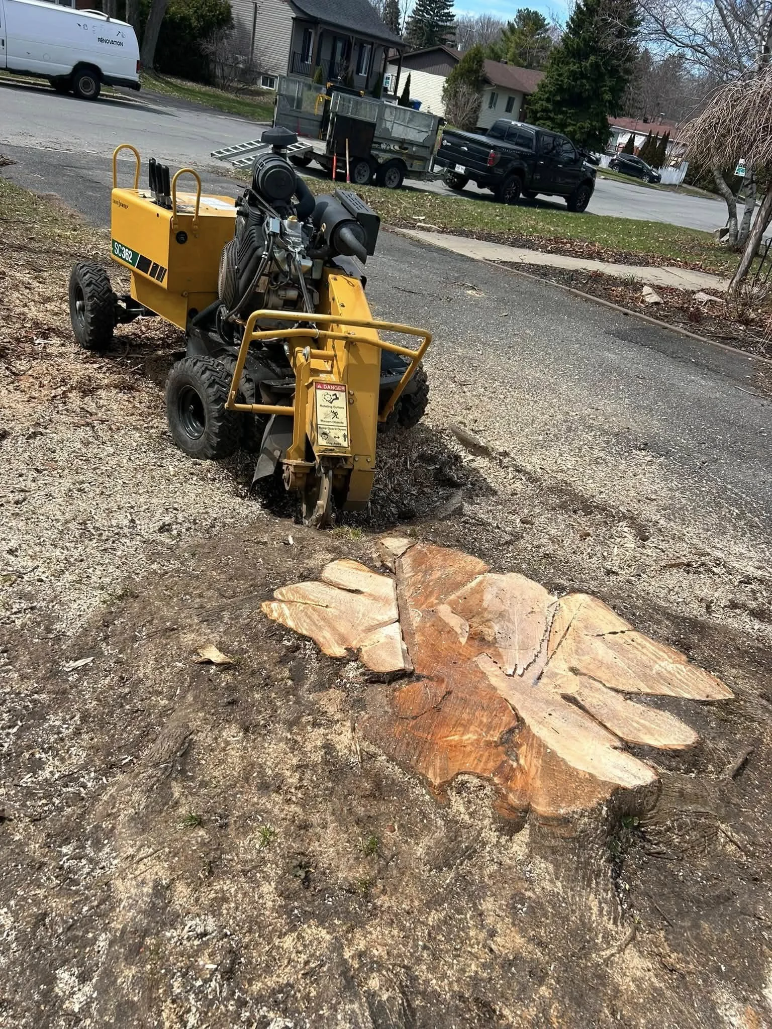 Une machine de coupe utilisée pour couper une branche d'arbre massive, située sur un sol en gravier. En arrière-plan, il y a des maisons résidentielles, des voitures et un remorque, dans une zone résidentielle.