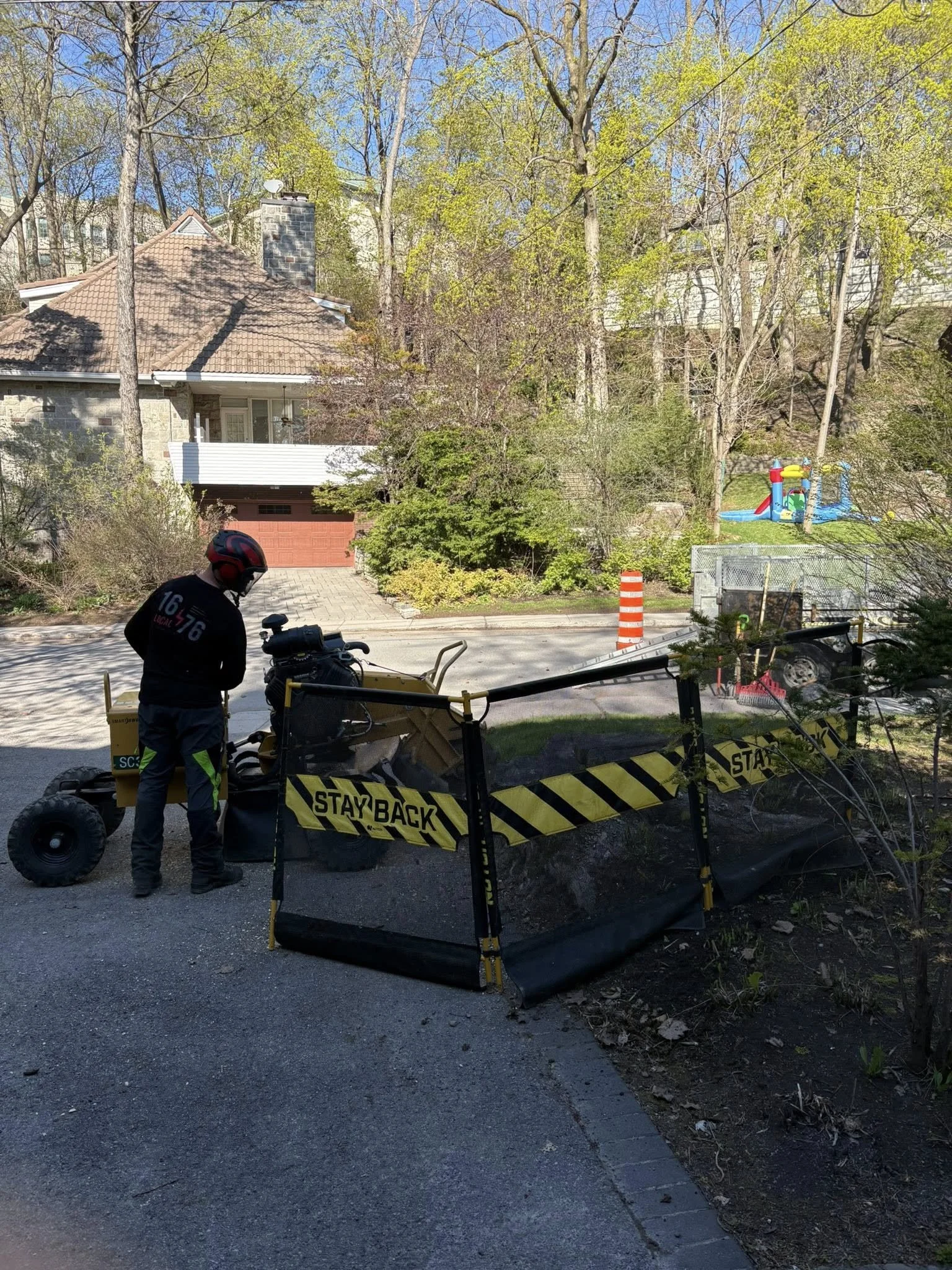 Travaux de voirie avec un panneau de signalisation « Restez à l'écart » en jaune et noir, un homme en casque noir et veste noire à côté d'une machine de compactage sur la chaussée, en arrière-plan une maison avec un balcon, des arbres feuillus et un espace de jeux pour enfants.