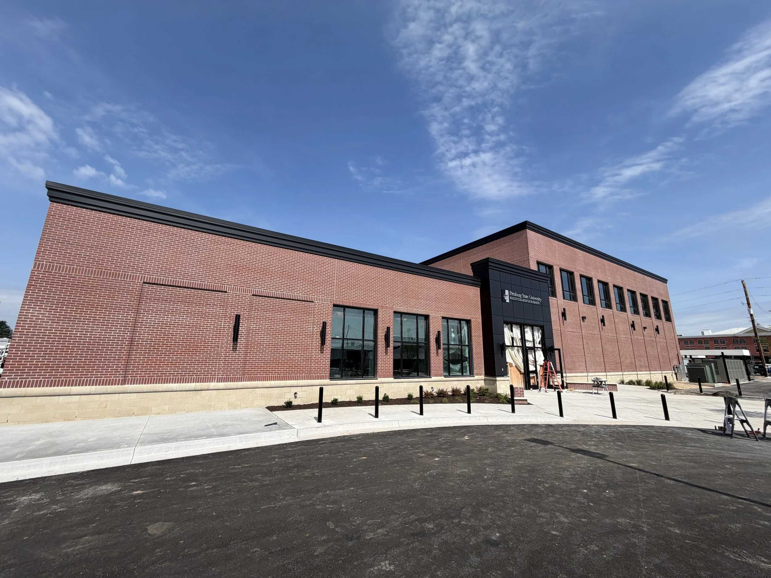 A modern brick building with large glass windows and a sign that reads 'Pittsburgh State University, College of Business' under a blue sky with scattered clouds.