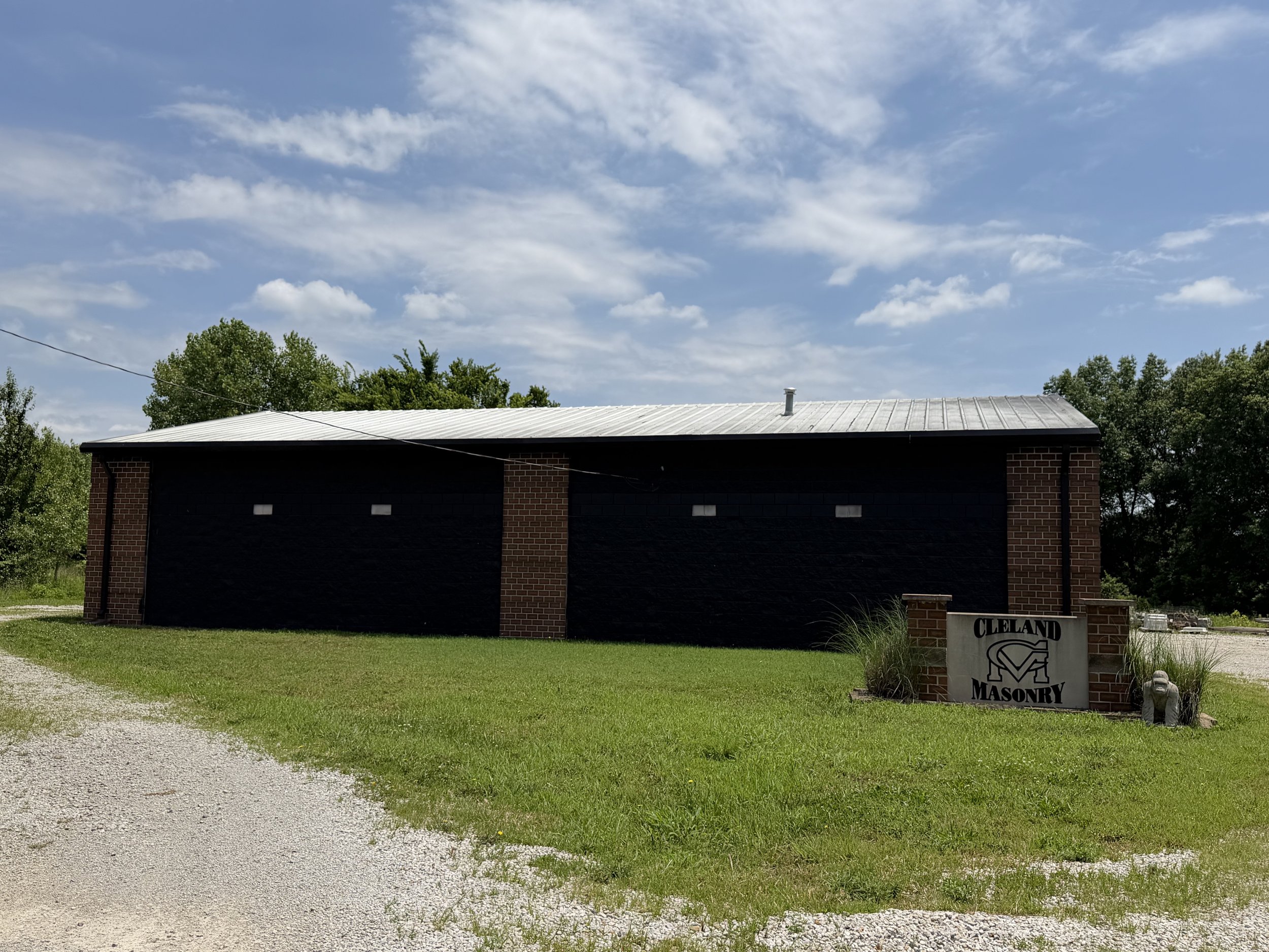 A building with a brick wall and metal roof, a sign that says 'Cleveland Masonry', green grass, trees, and a blue sky with some clouds.