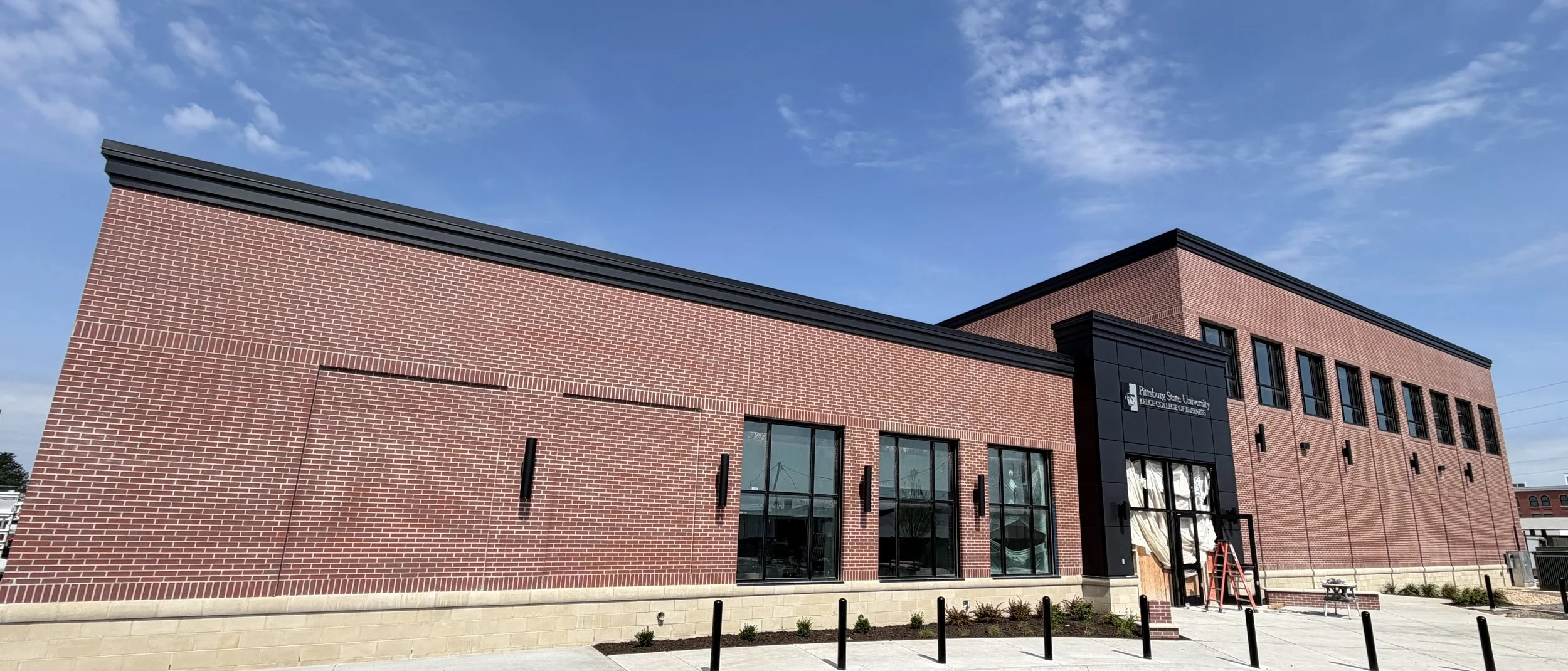 A modern brick building with large glass windows and a sign that reads 'Pittsburgh State University, College of Business' under a blue sky with scattered clouds.