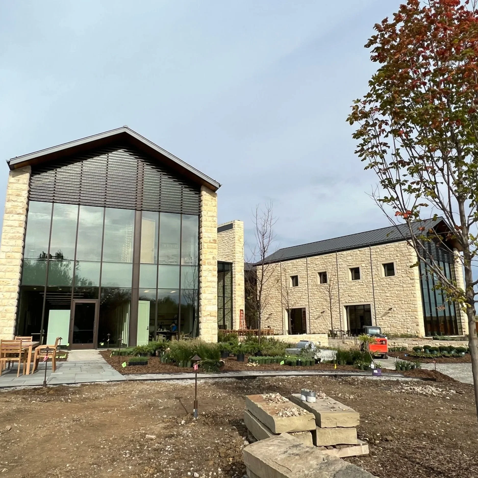 Modern building under construction with a large glass facade, surrounded by a garden with wooden chairs and planting areas, a tree with red and green leaves, and construction materials on the ground.