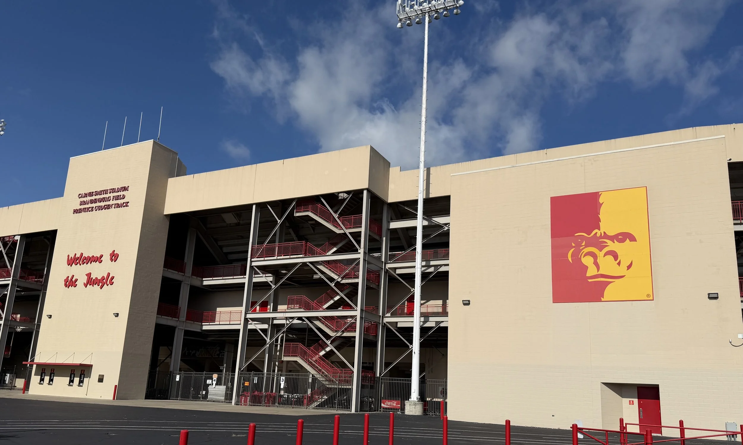 Exterior view of a large stadium with a beige facade, red staircases, and a prominent lion logo. Signage indicating it is the Cal State San Bernardino Stadium, Branduburg Field, and the Precision Cutter Track, with a parking area and red safety posts in the foreground under a blue sky with clouds.