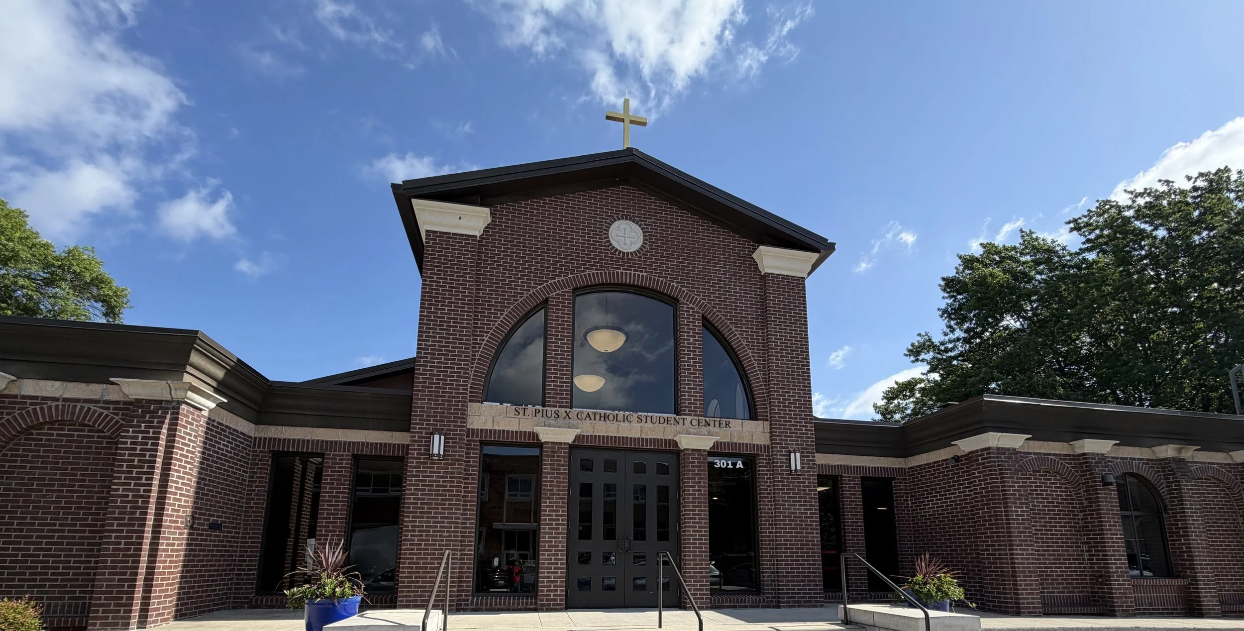 Brick church building with a cross on the roof, large arched window, and sign reading 'St. Pius X Catholic Student Center' under a blue sky with clouds and trees on the sides.
