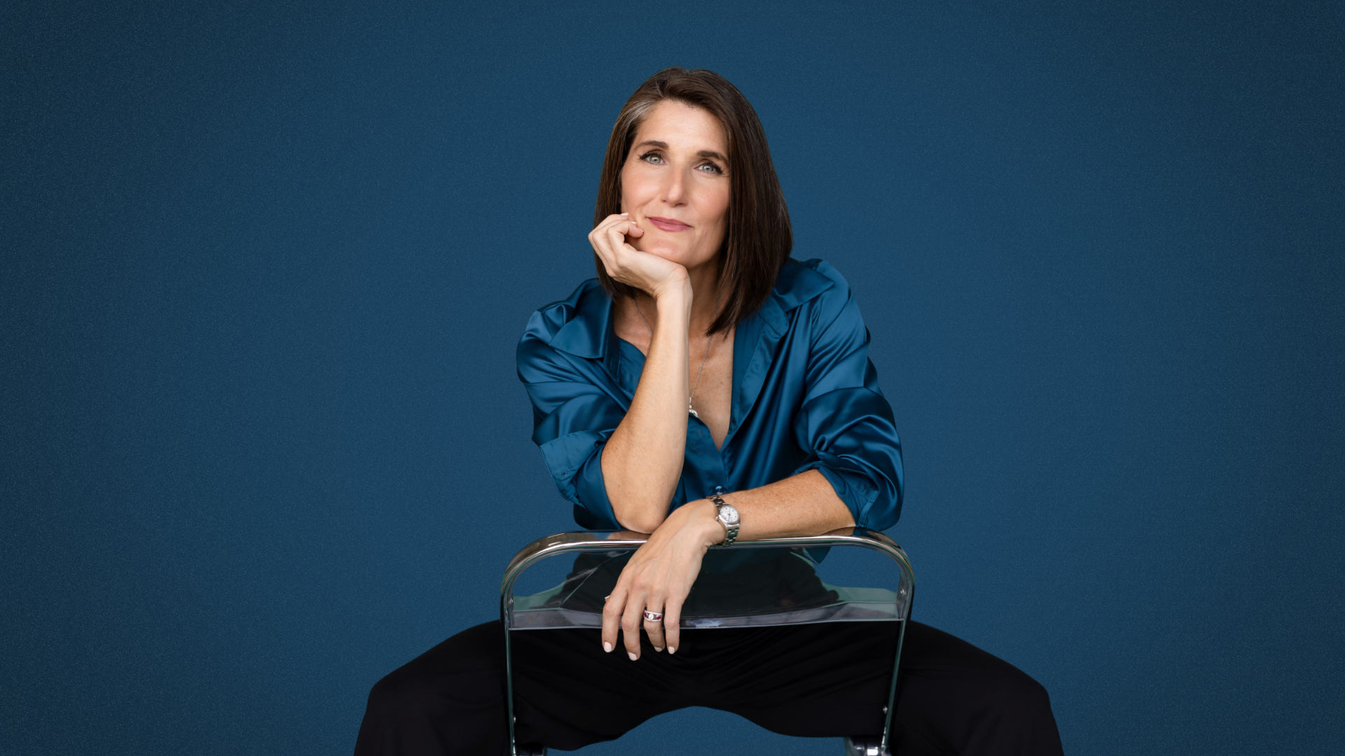 A woman with brown hair wearing a blue satin blouse sitting on a transparent chair against a dark blue background, resting her chin on her hand and looking at the camera.