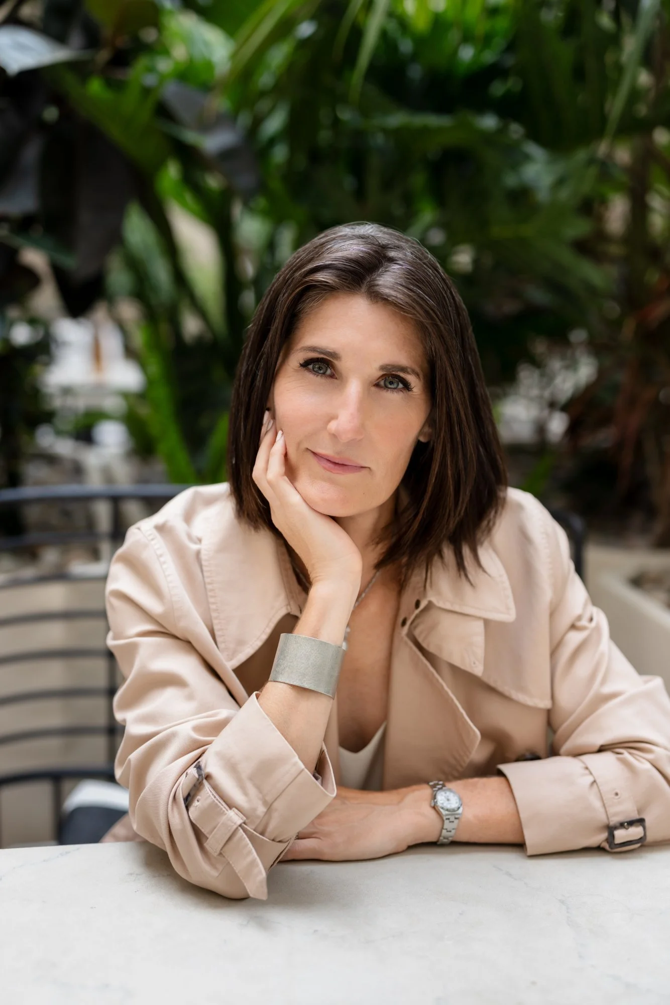 Woman with brown hair and blue eyes resting her chin on her hand, sitting at a table with a green, leafy background.
