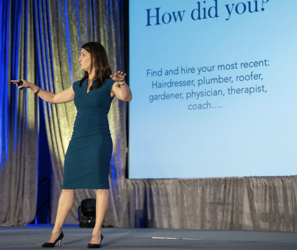 A woman in a teal dress giving a presentation on stage with a large screen behind her displaying text about hiring various professionals.