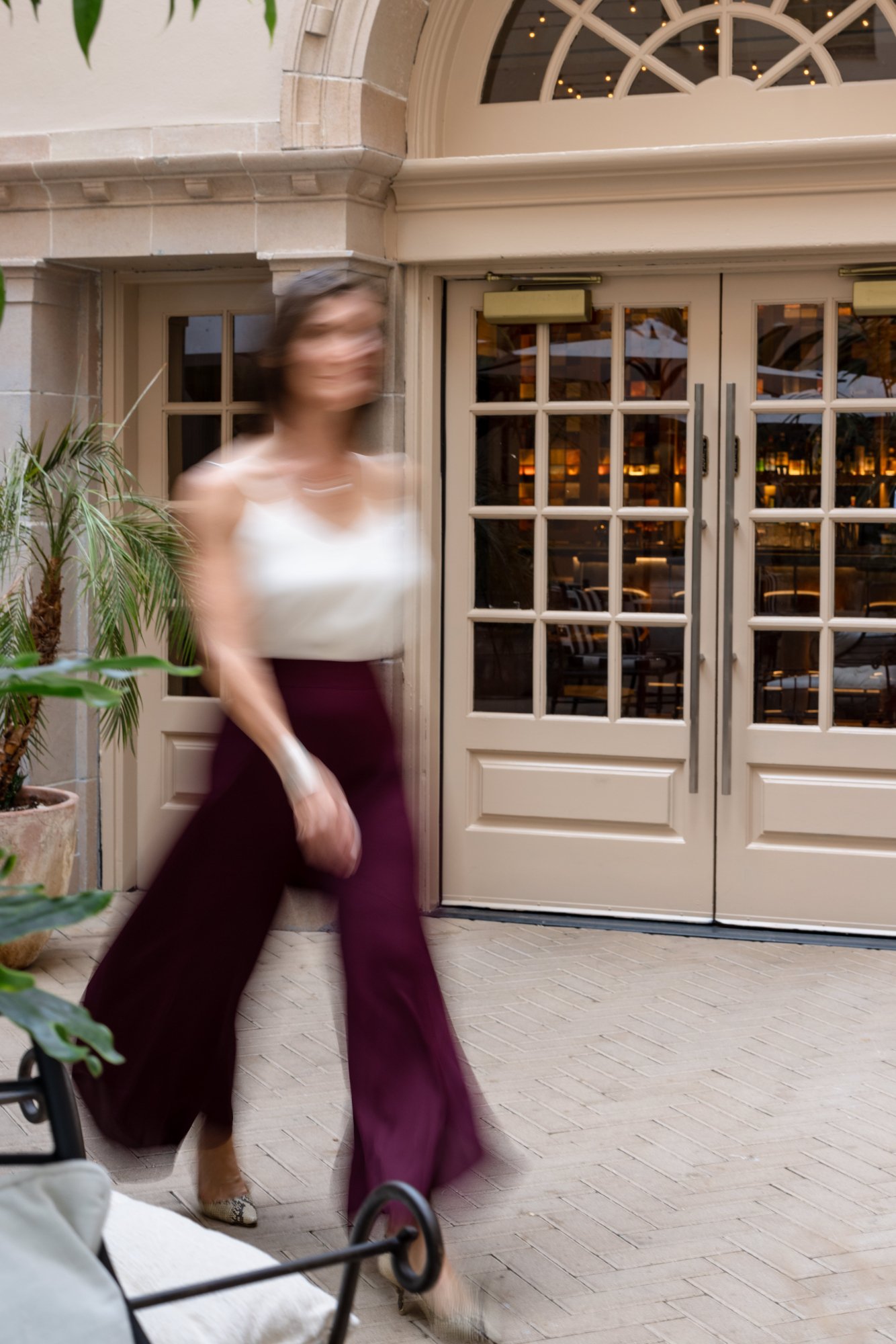 A woman walking past a door with glass panels inside a building or restaurant, with plants nearby and some furniture visible through the glass.