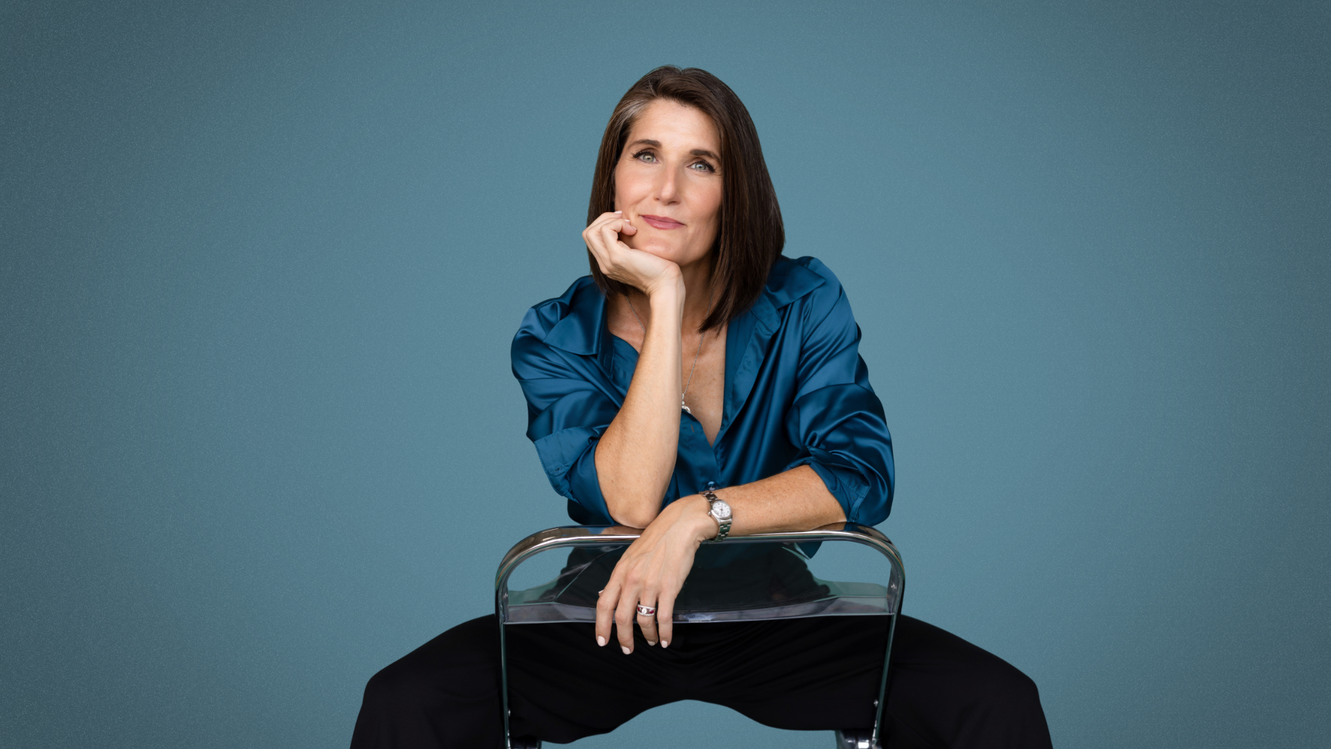 A woman with shoulder-length brown hair wearing a blue satin blouse, seated and resting her chin on her left hand, looking at the camera against a teal background.