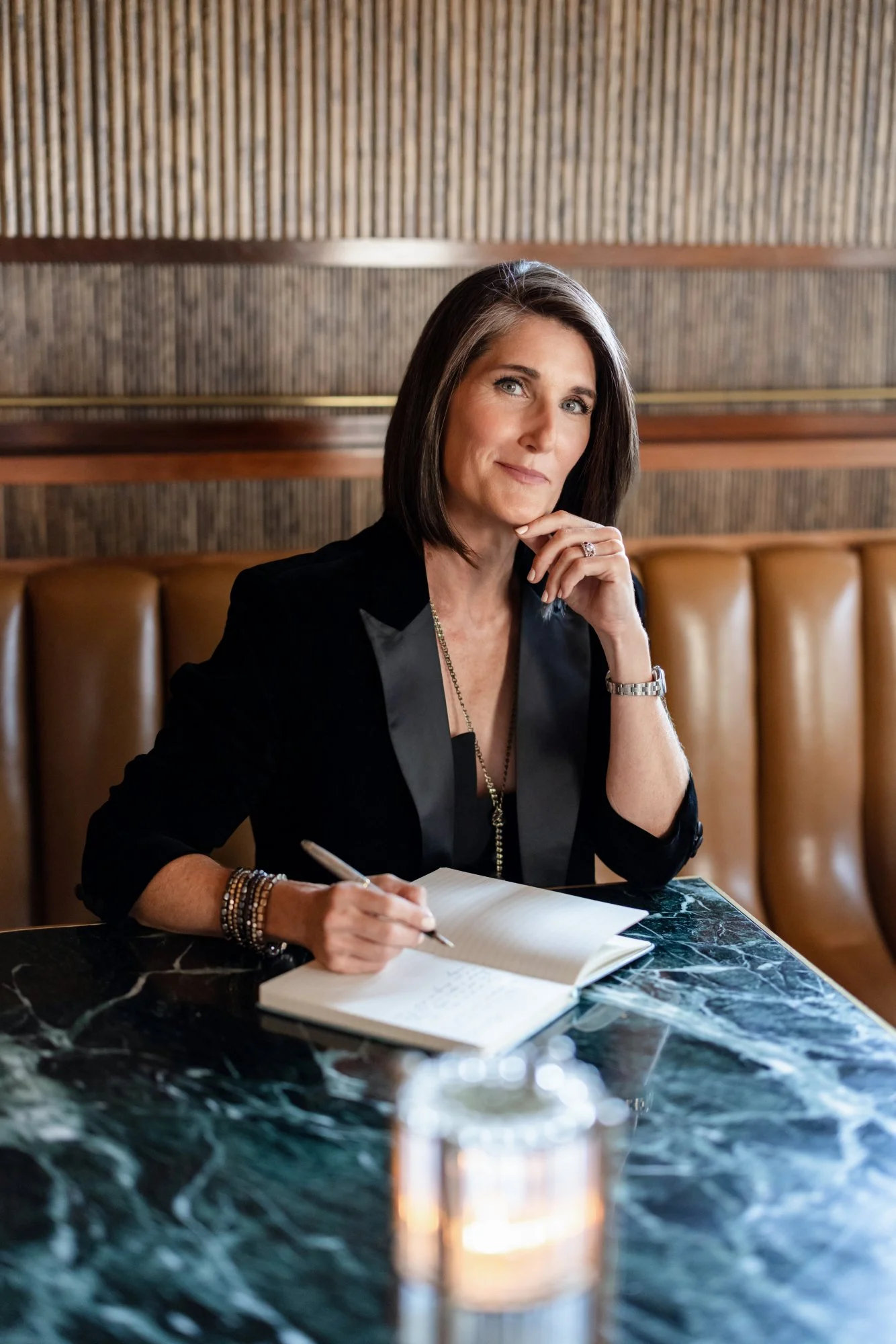 A woman with shoulder-length dark hair sitting at a black marble table, holding a pen and writing in a notebook. She is wearing a black blazer, a silver watch, and multiple bracelets. The background features wood-paneled walls and brown leather seating.