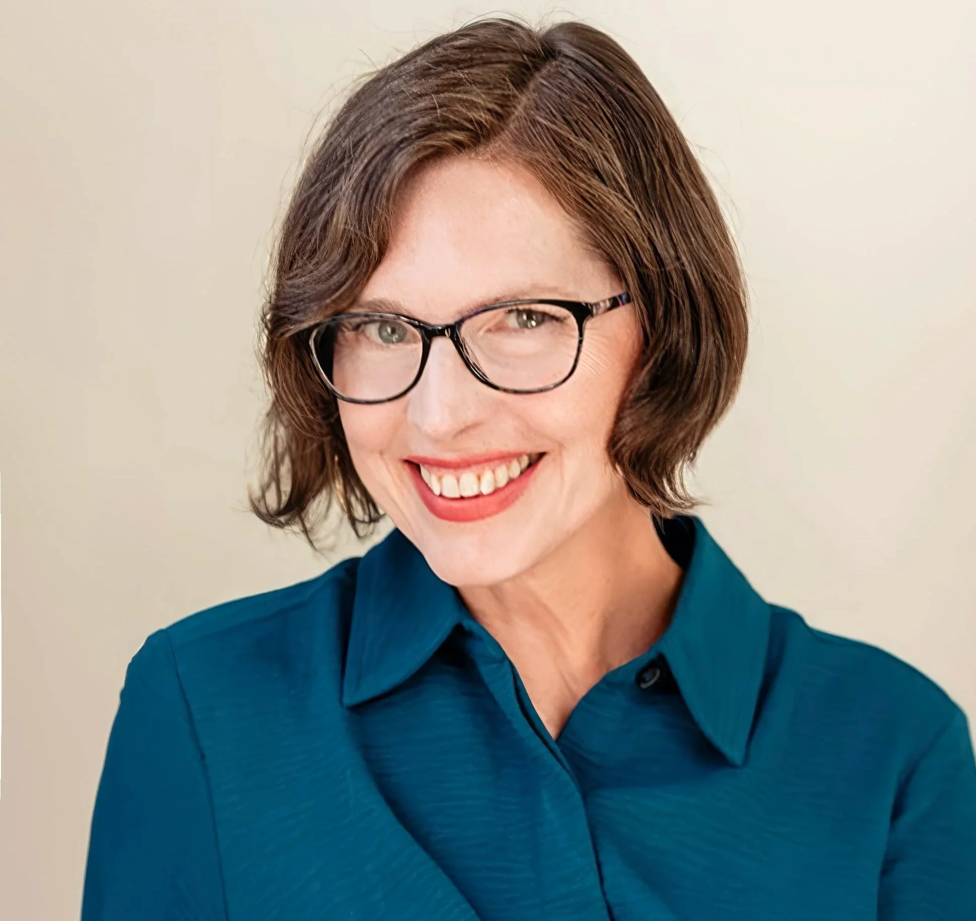 Close-up photo of a smiling woman with short brown hair, wearing glasses and a teal shirt against a plain light-colored background.