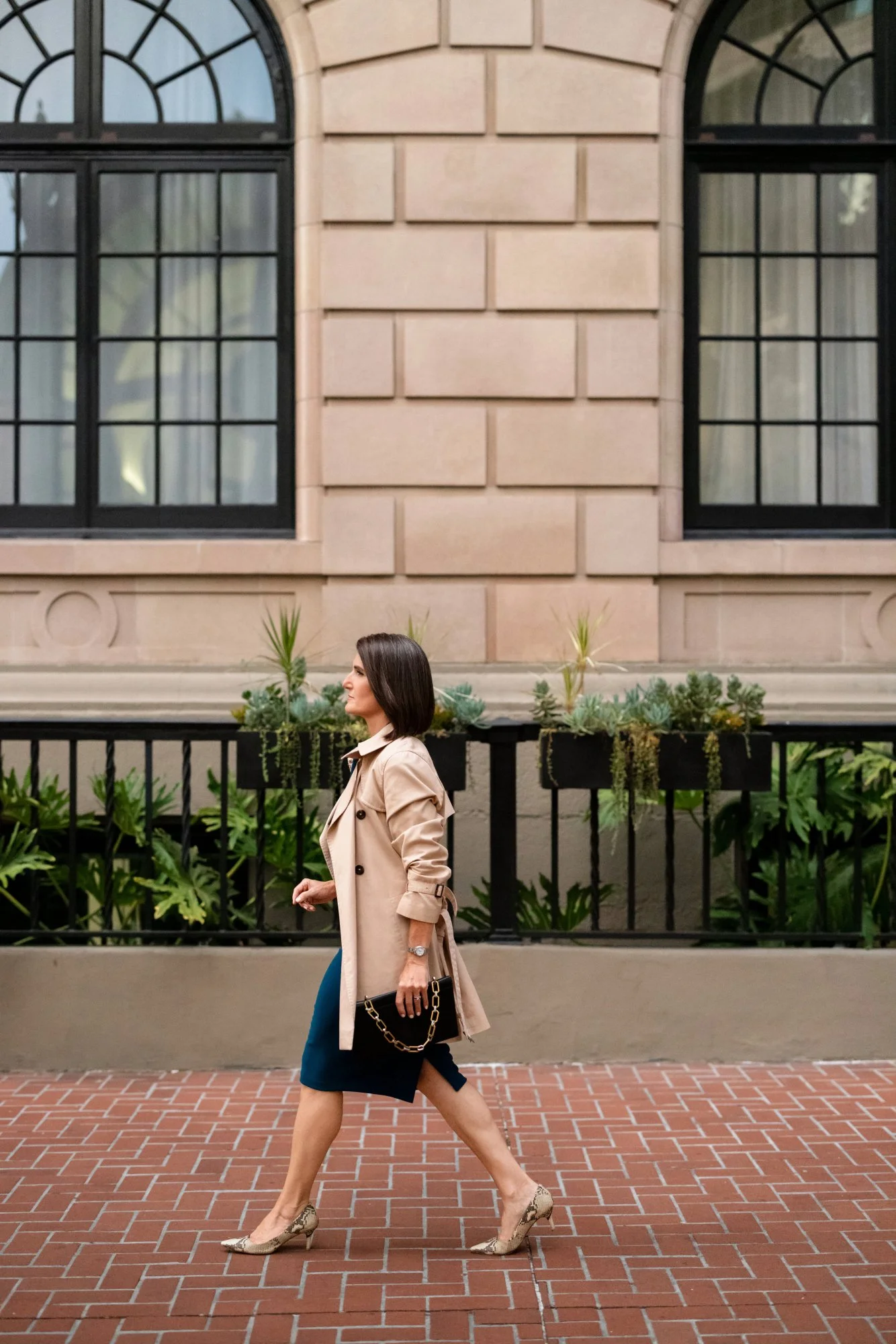 Woman walking on a brick sidewalk in front of a building with large arched windows and a decorative garden bed with green plants.