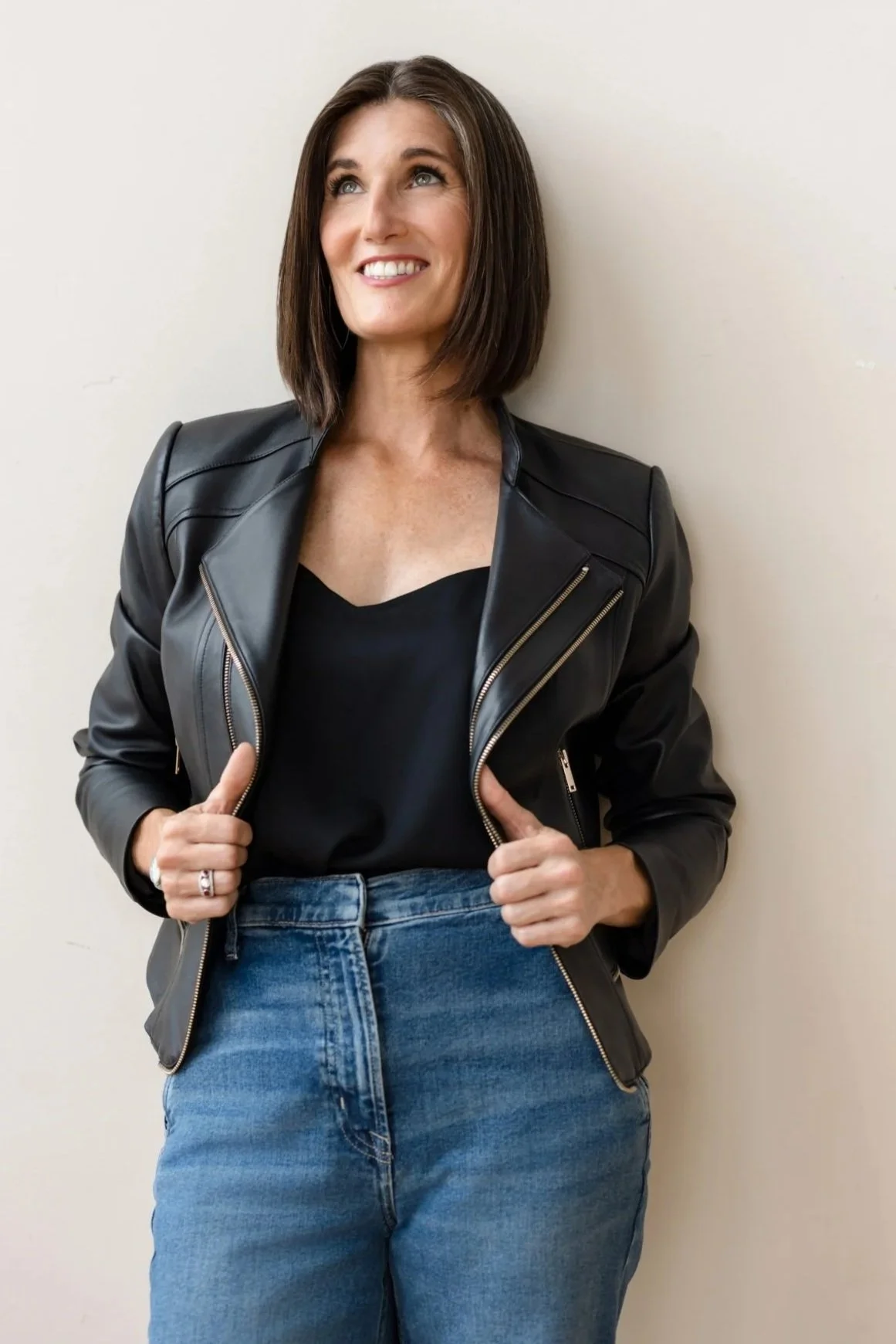 Woman with shoulder-length dark hair smiling, wearing a black leather jacket over a black top and blue jeans, standing against a plain white wall.