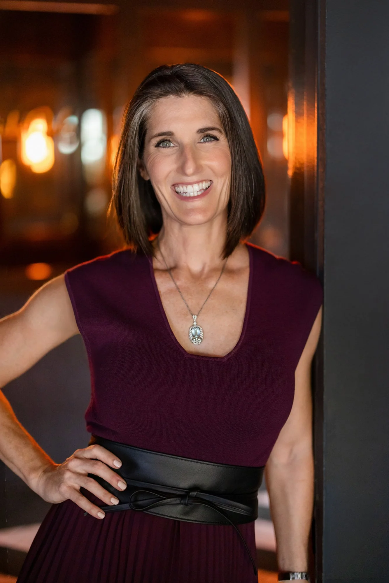 A smiling woman with shoulder-length brown hair, wearing a sleeveless maroon top, a silver pendant necklace, and a watch, standing indoors near a dark wall with warm bokeh lighting in the background.