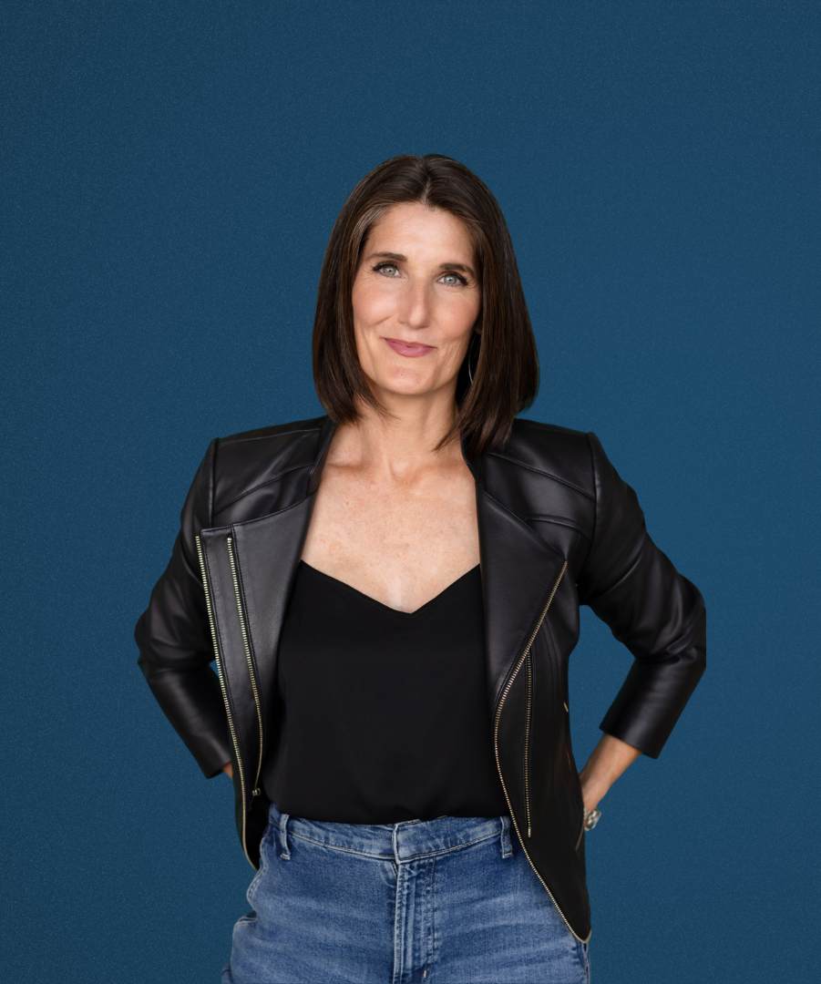 A woman with shoulder-length brown hair, wearing a black leather jacket over a black top and blue jeans, stands against a blue background, smiling slightly with her hands behind her back.