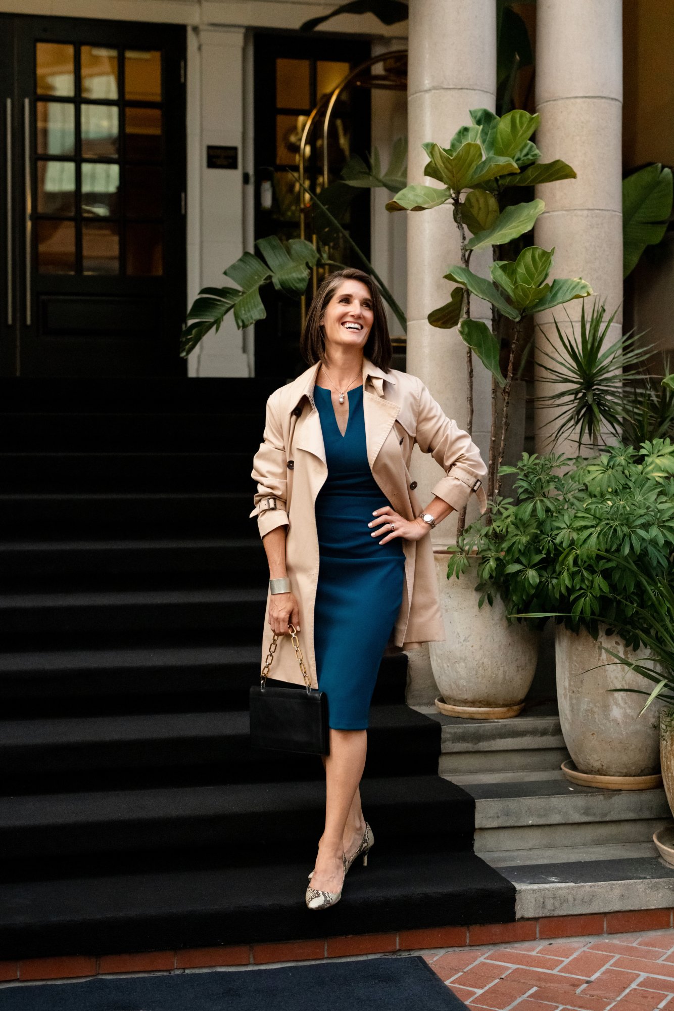 Woman standing on black stairs outside a building, smiling, wearing a beige trench coat over a royal blue dress, holding a black purse with a gold chain, surrounded by large potted plants and columns.