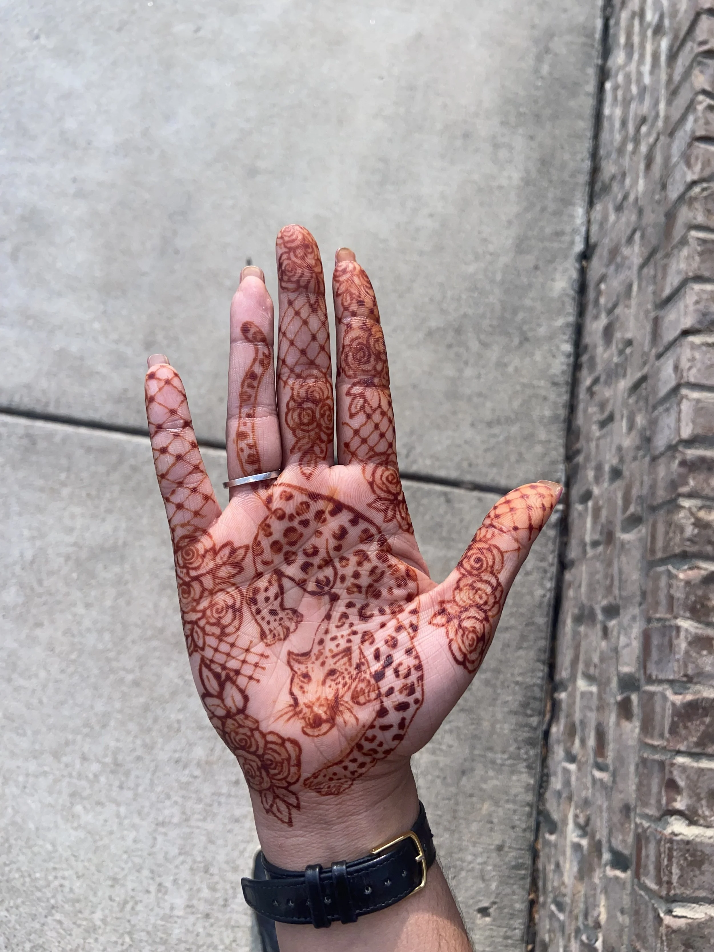 A person's hand decorated with intricate henna designs, with a brick wall and sidewalk in the background.