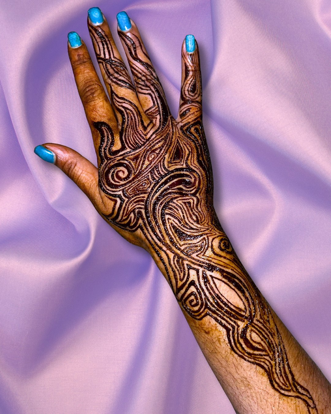 A hand with intricate black henna designs on the palm and fingers, and blue nail polish, resting on a soft pink fabric background.