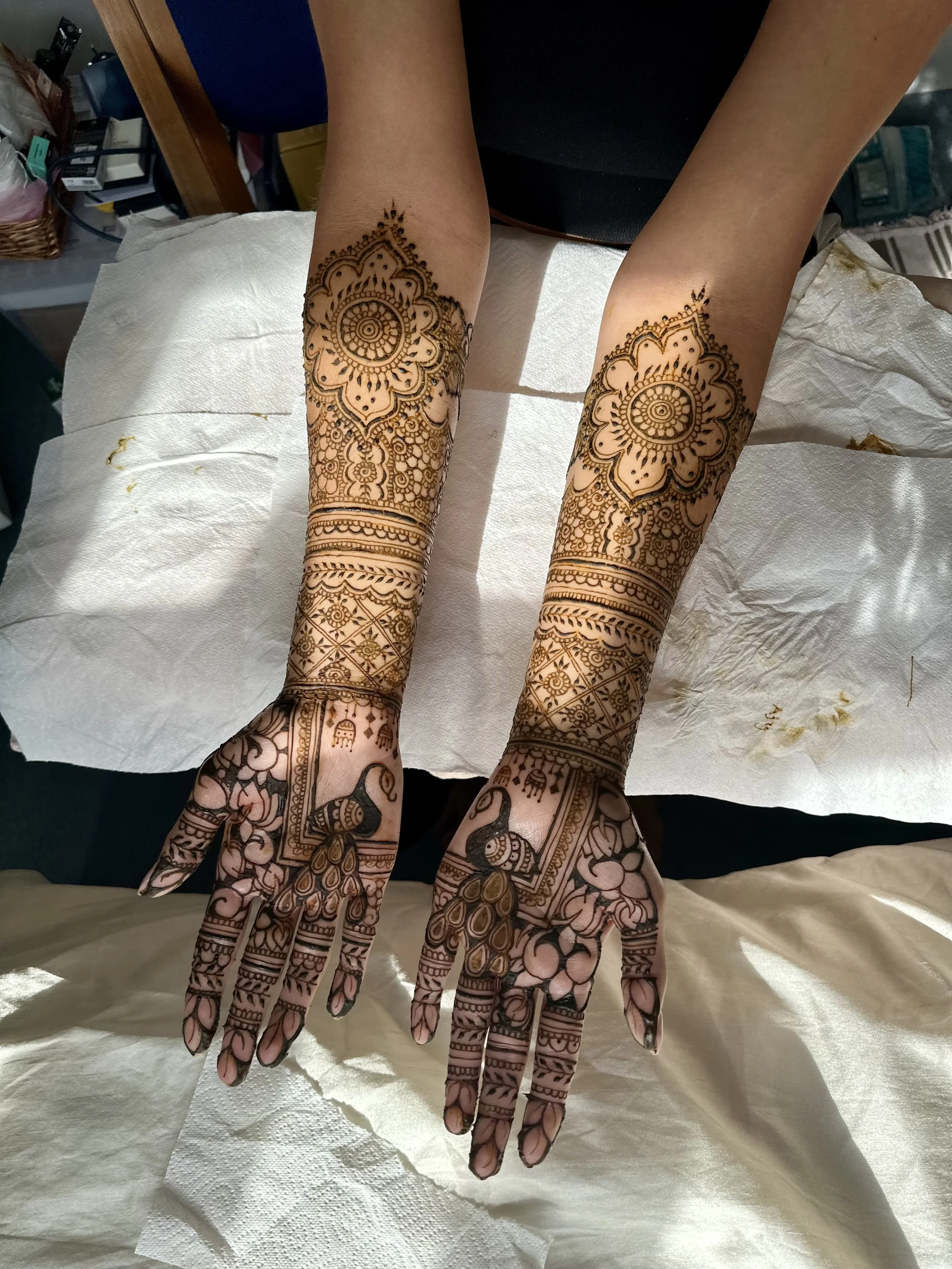 Person demonstrating intricate henna tattoo designs on both hands and forearms, with detailed patterns including floral and geometric motifs.