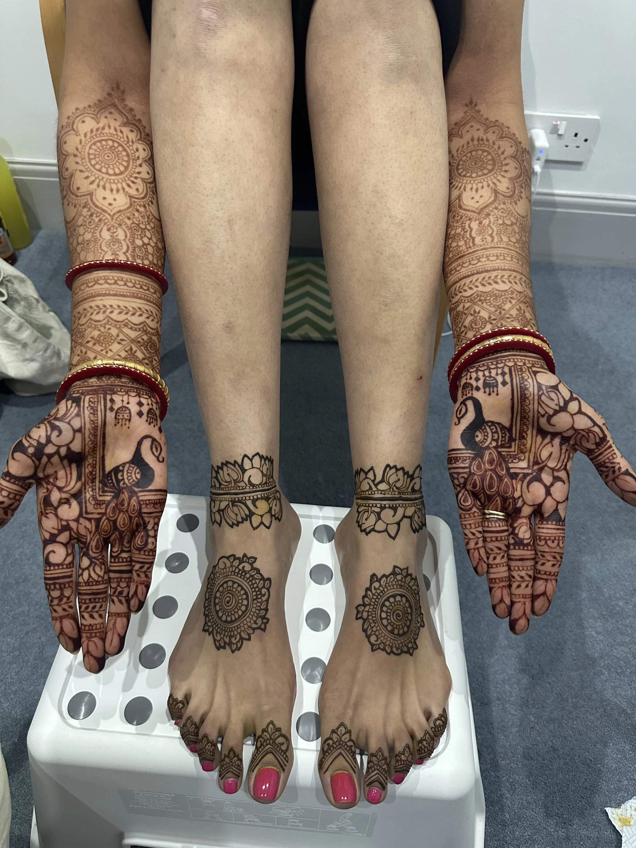 Hands and feet decorated with intricate henna designs resting on a white surface, with pink nail polish on fingernails and toenails, and wearing matching red and gold bangles.