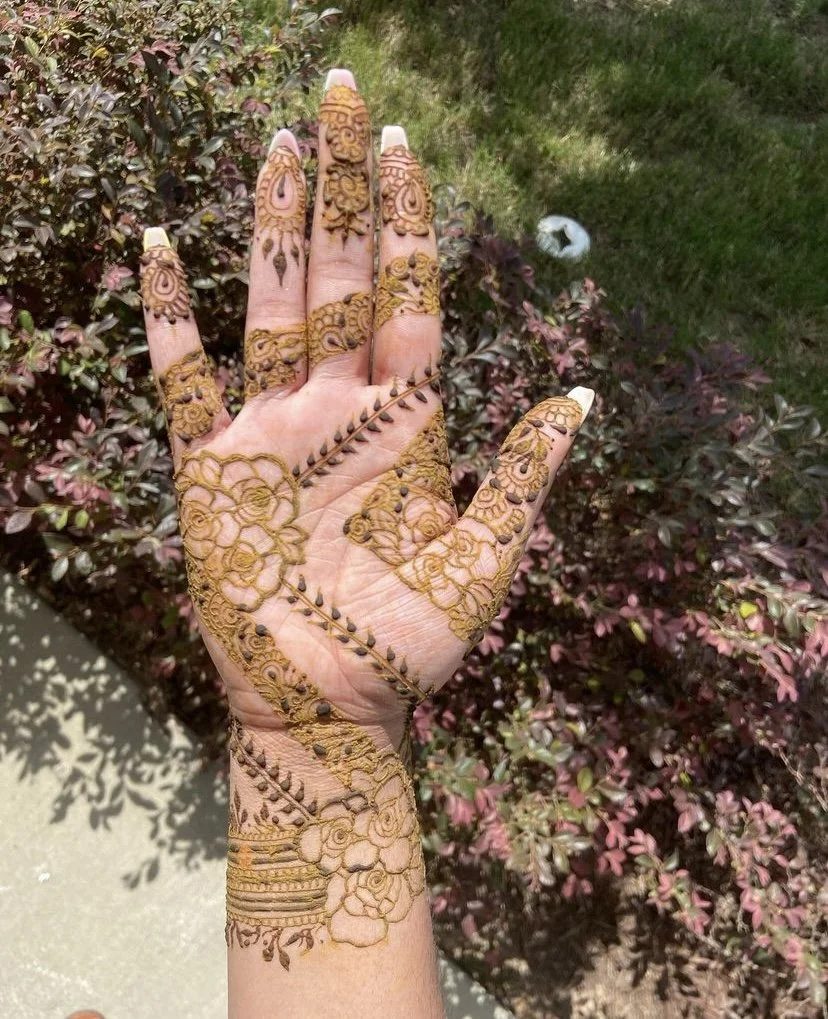 Hand decorated with intricate henna designs featuring floral and geometric patterns, held against a background of green and purple shrubs.