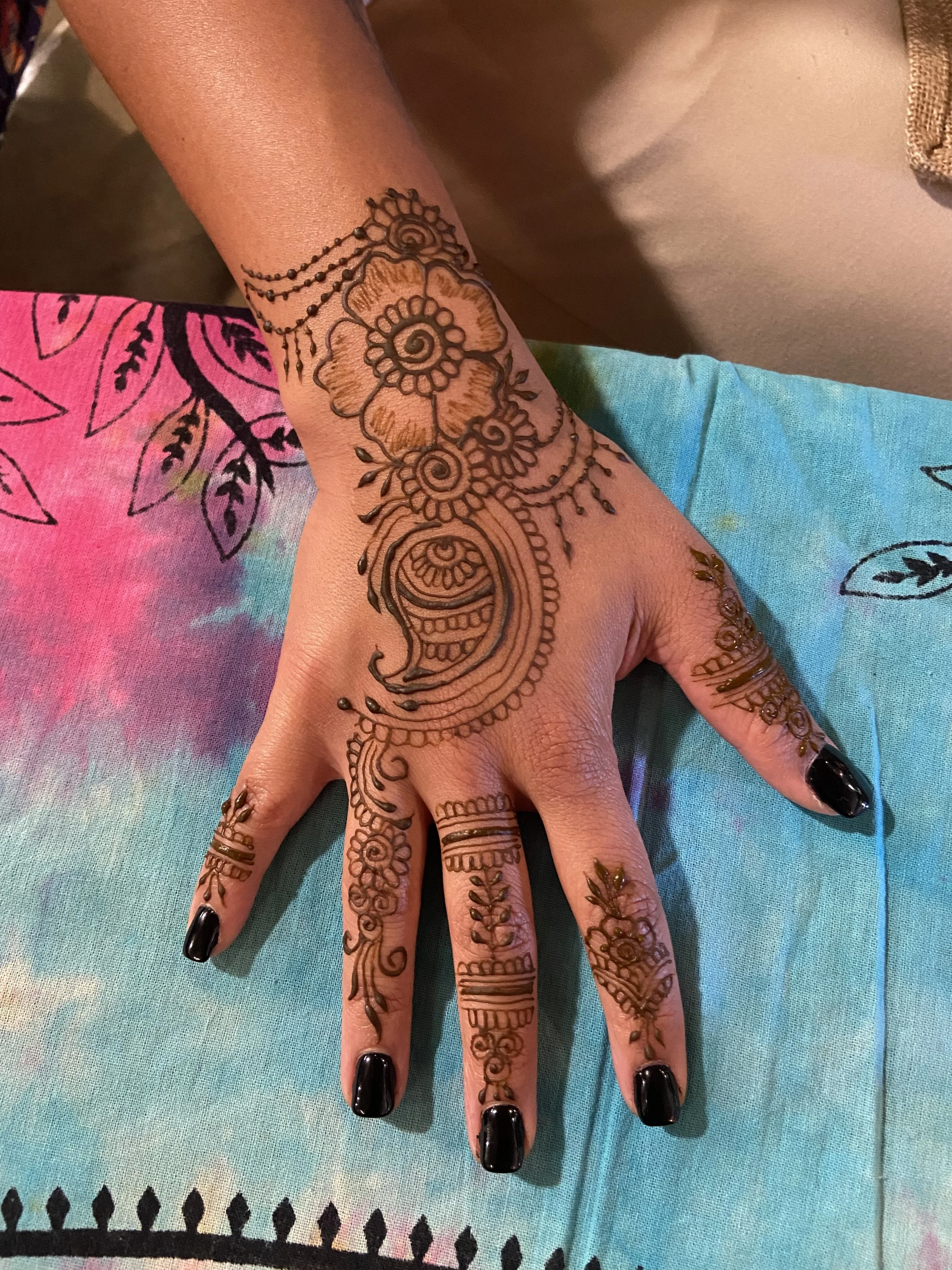 Hand with intricate henna designs on fingers, palm, and wrist, with black nail polish, resting on fabric with tie-dye rainbow colors and black geometric patterns.