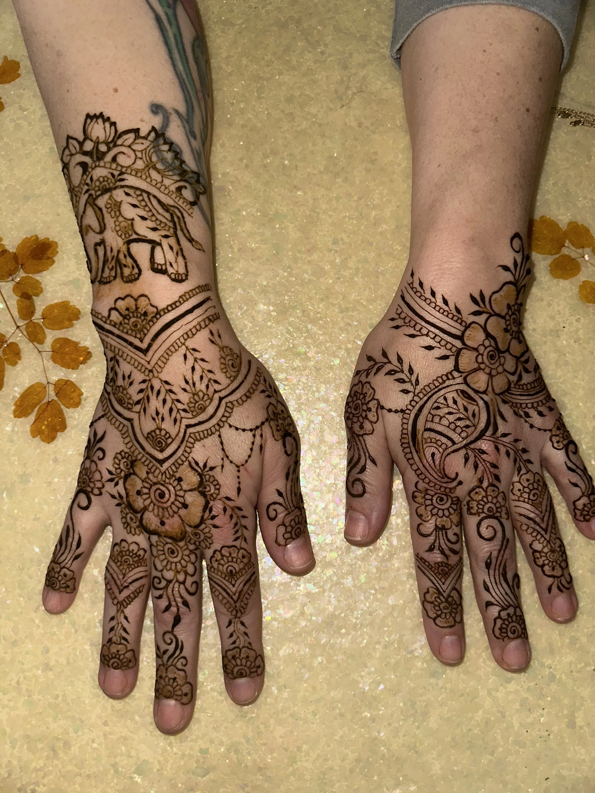 Two hands with henna tattoos on fingers and back of hands, resting on a glittery yellow surface with small orange leaves nearby.