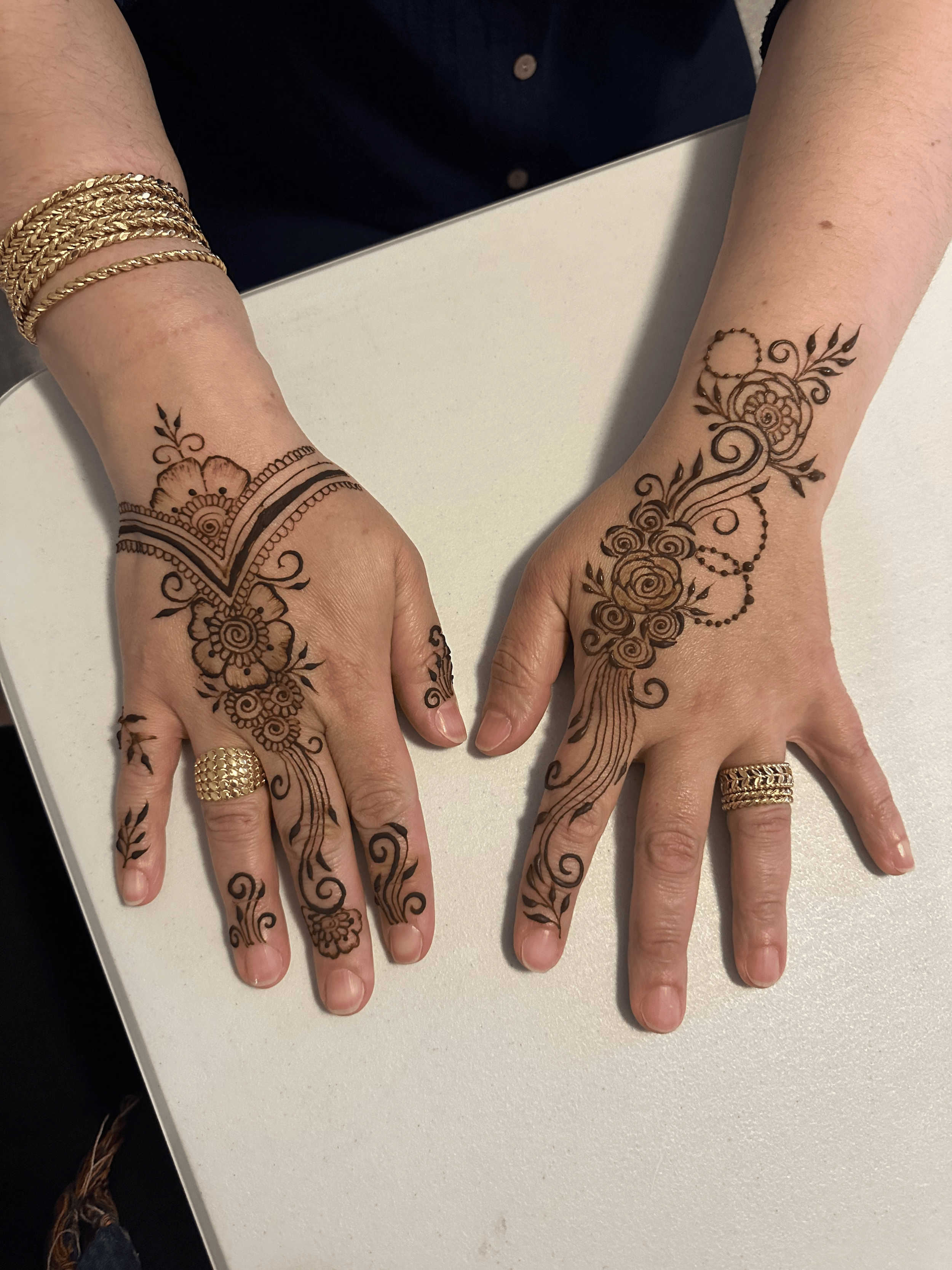 Two hands with henna designs on the fingers and back of the hands, wearing gold rings and bracelets, resting on a white surface.