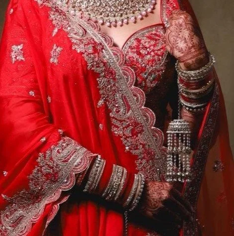 Close-up of a woman dressed in a red Indian bridal outfit with silver embroidery, wearing traditional jewelry, bangles, and mehndi designs on her hands, holding a mirror.