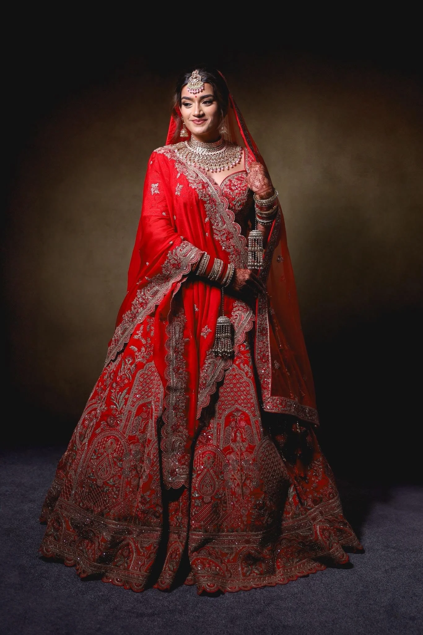 An Indian bride dressed in a red traditional bridal outfit, adorned with gold embroidery, jewelry, and henna on her hands.