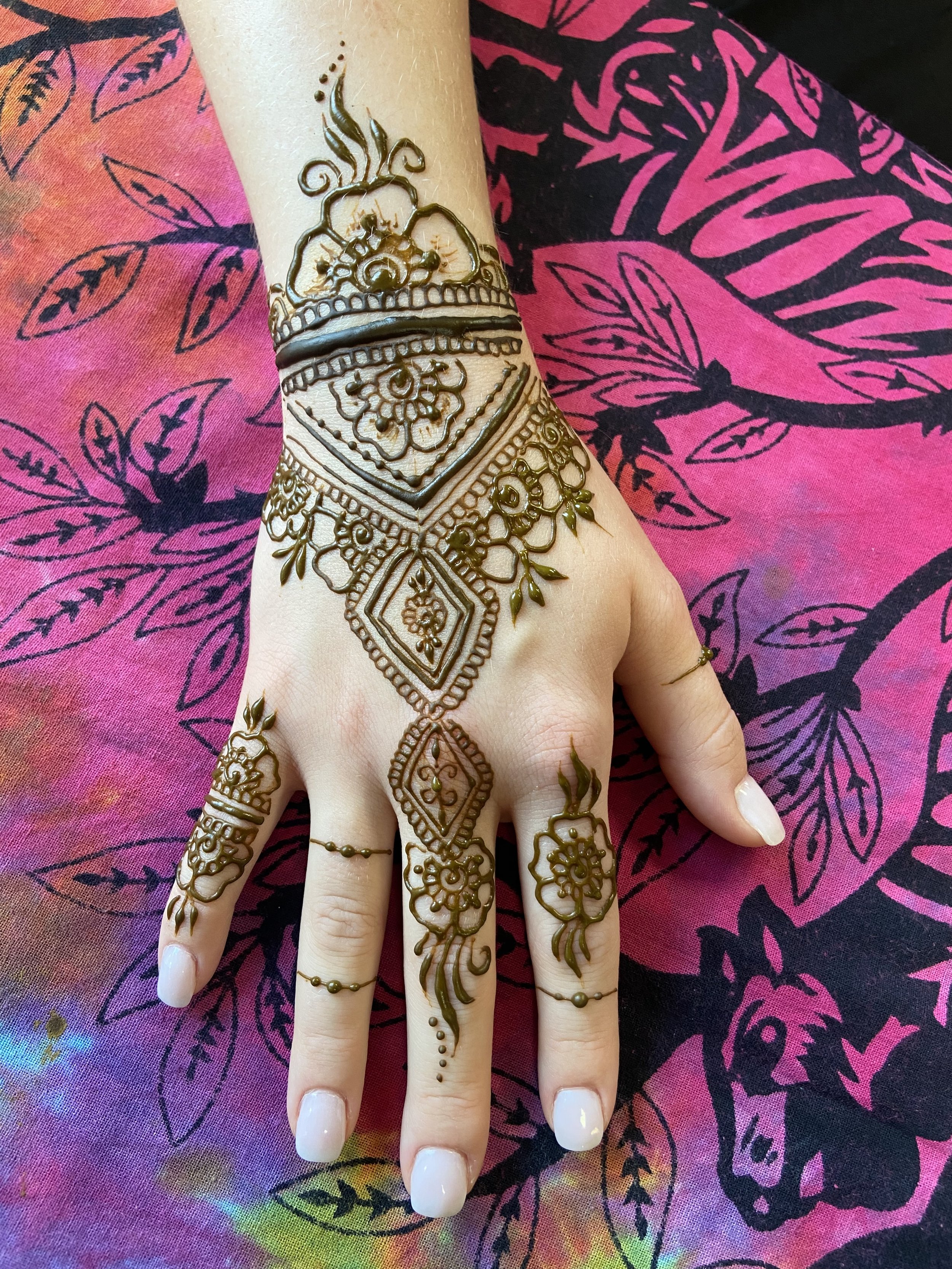 Close-up of a hand adorned with mehndi (henna) designs, resting on a colorful fabric with leaf and floral patterns.
