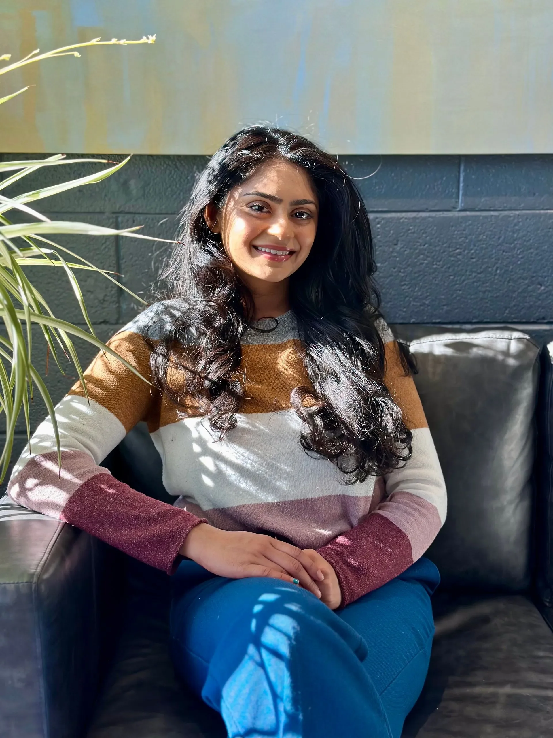 A woman with long black curly hair and a warm smile sitting on a black leather couch near a blue wall with colorful painting and a green plant leaves in the foreground.