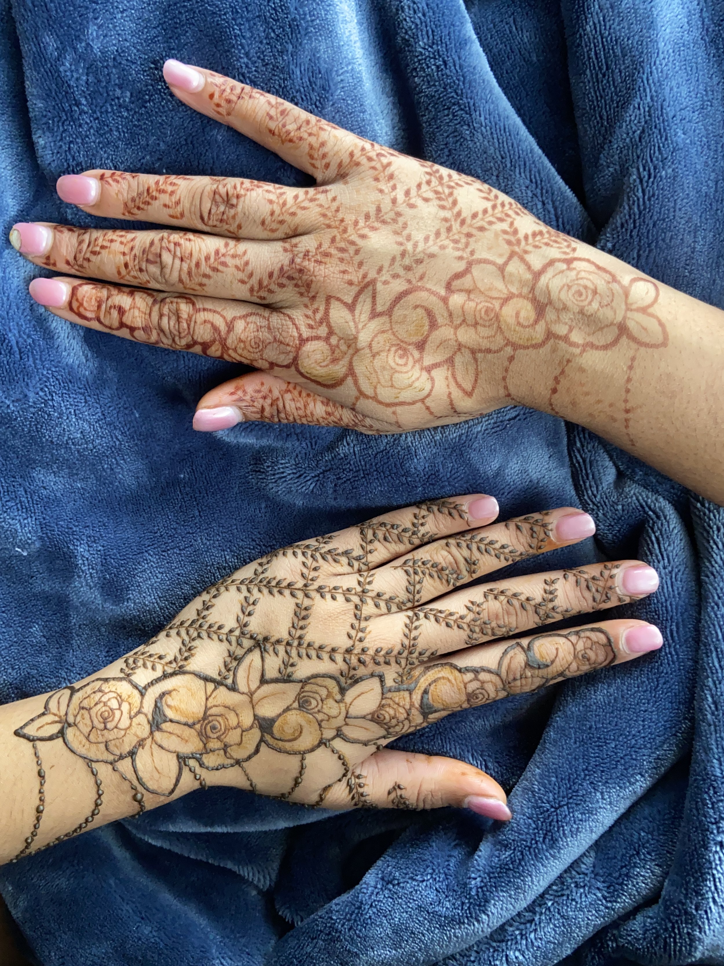 Close-up of two hands with intricate henna designs on fingers, palms, and wrists, resting on a blue fabric background.
