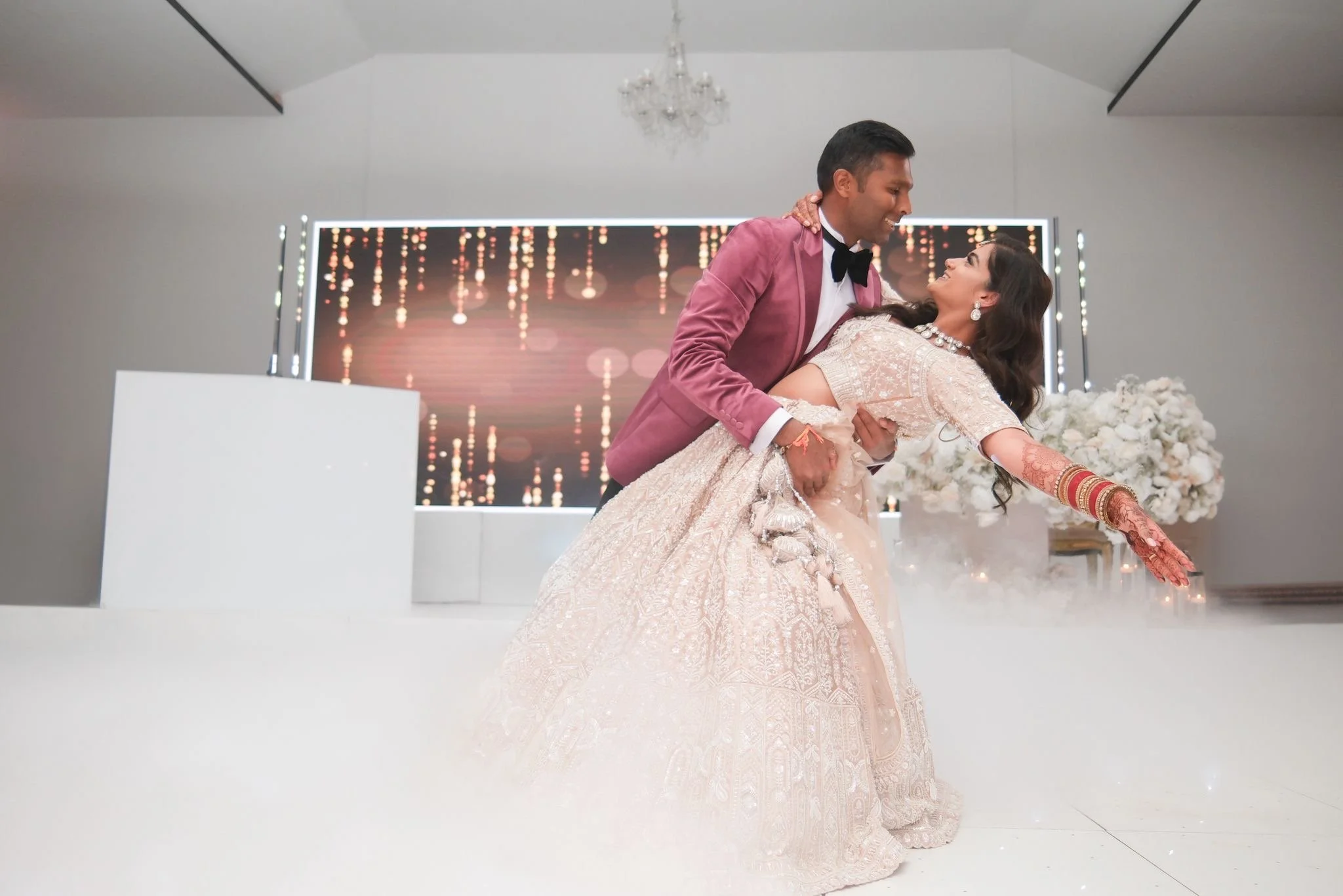 A bride and groom dancing at their wedding reception. The groom is wearing a coral pink tuxedo with a black bow tie, and the bride is dressed in a cream-colored traditional gown with intricate embroidery, jewelry, and mehndi on her arm. The bride is leaning back as they dance, smiling at each other. The background features a digital display with bokeh light effects, white flowers, and a chandelier hanging from the ceiling.