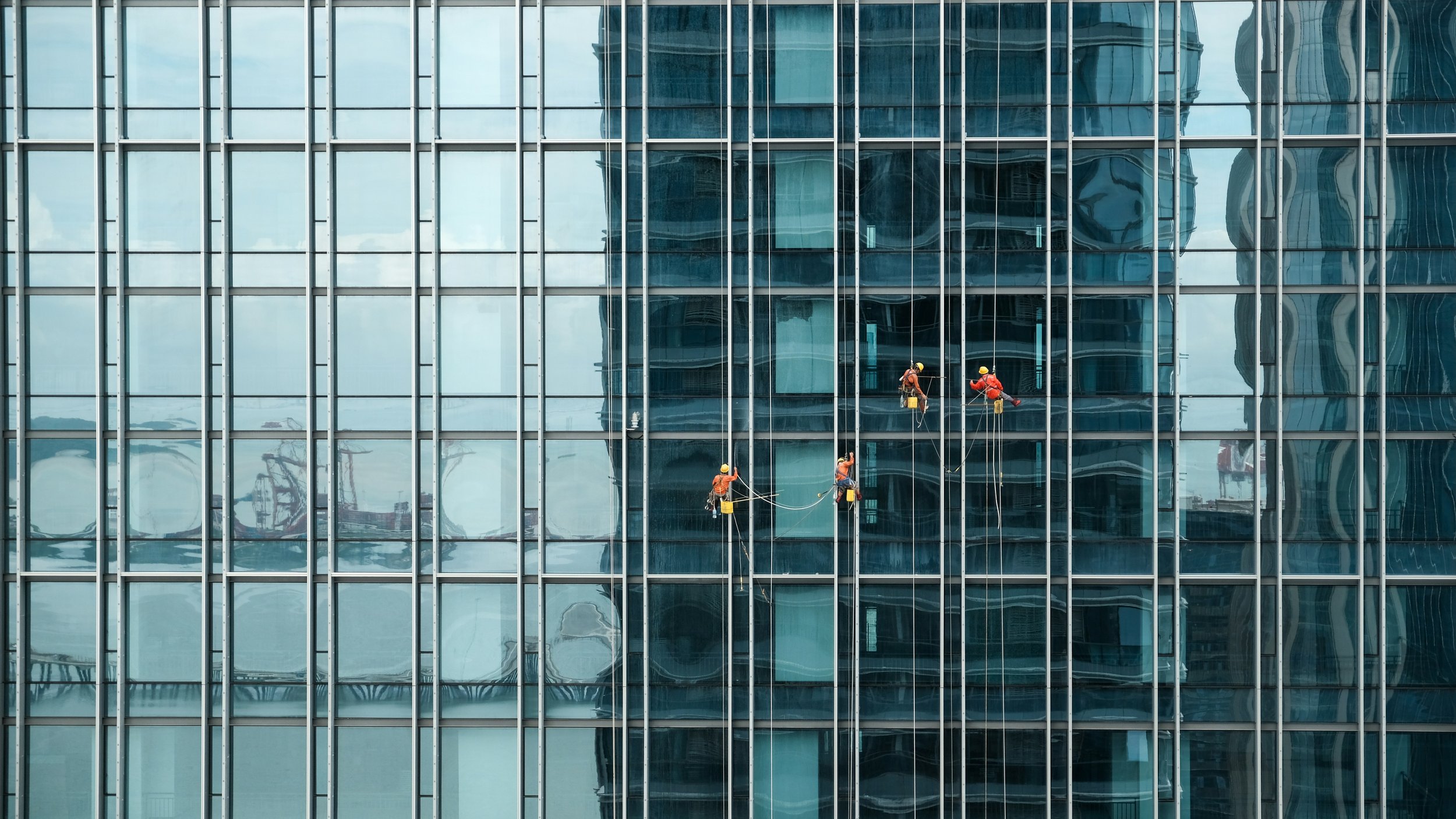 Workers in safety gear cleaning the exterior windows of a tall glass building.