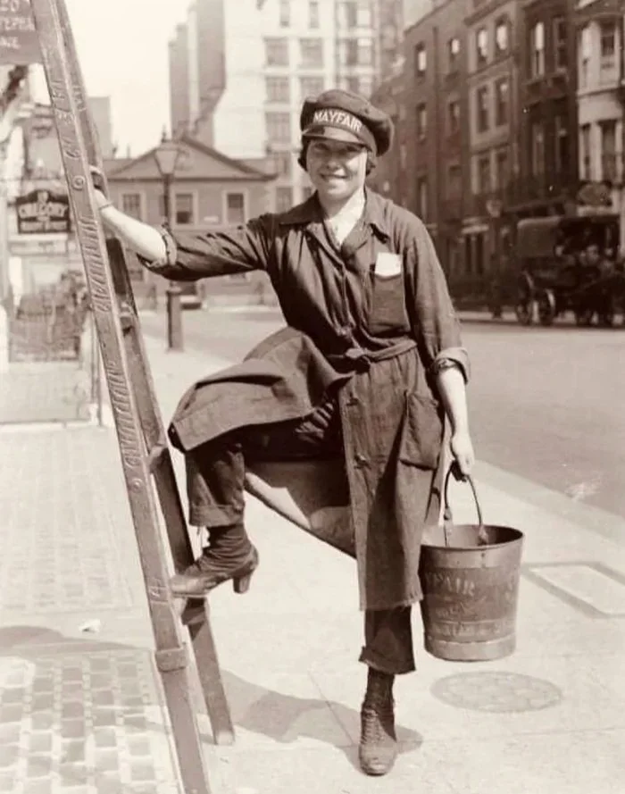 Vintage photograph of a smiling young woman with a piece of cloth on her head, standing on a city sidewalk next to a stepladder, holding a bucket, with city buildings and a horse-drawn carriage in the background.
