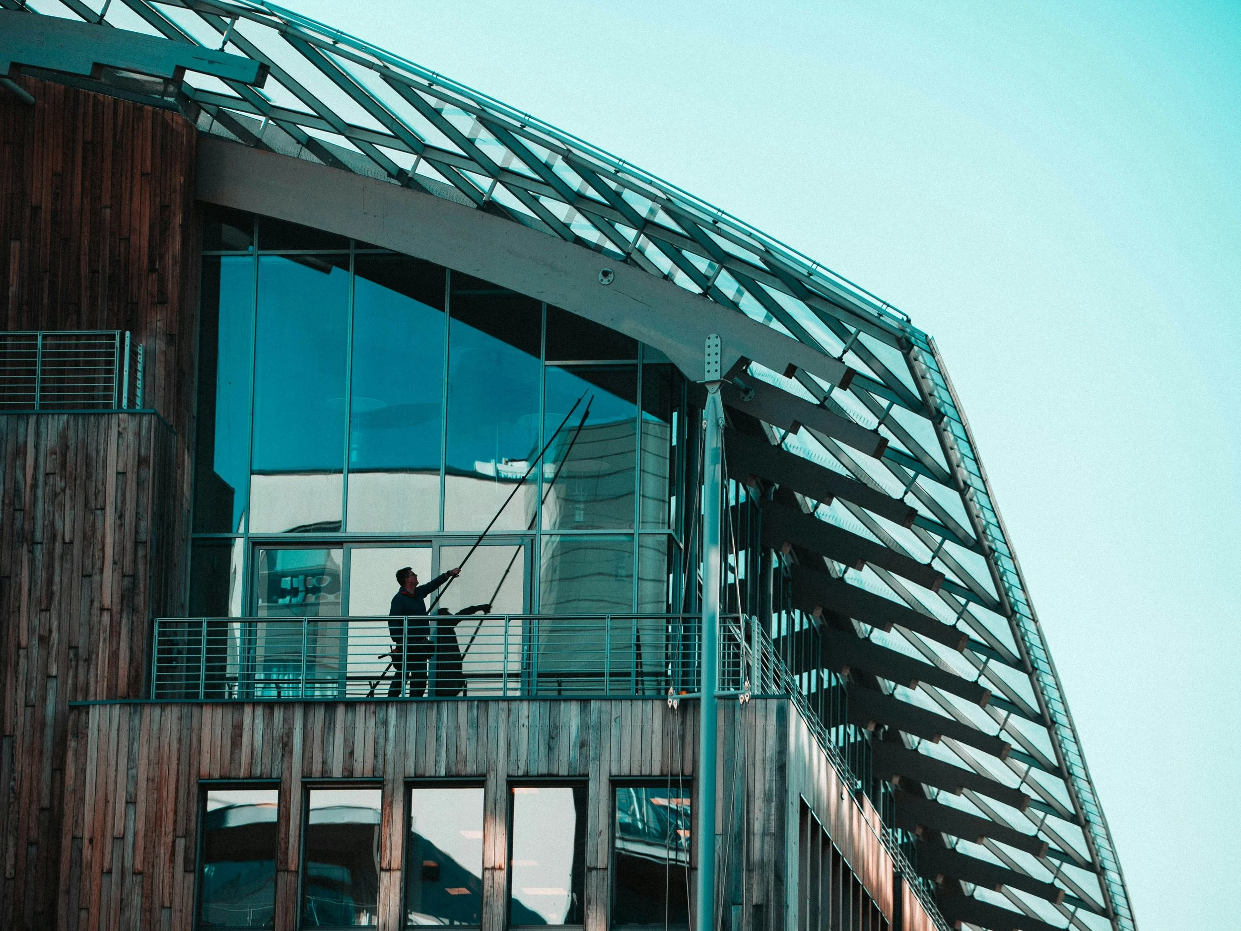 A man on a balcony cleaning the glass windows of a modern building with a long pole. The building features a combination of wood paneling, glass, and unique architectural elements, including a curved glass structure on the upper part.