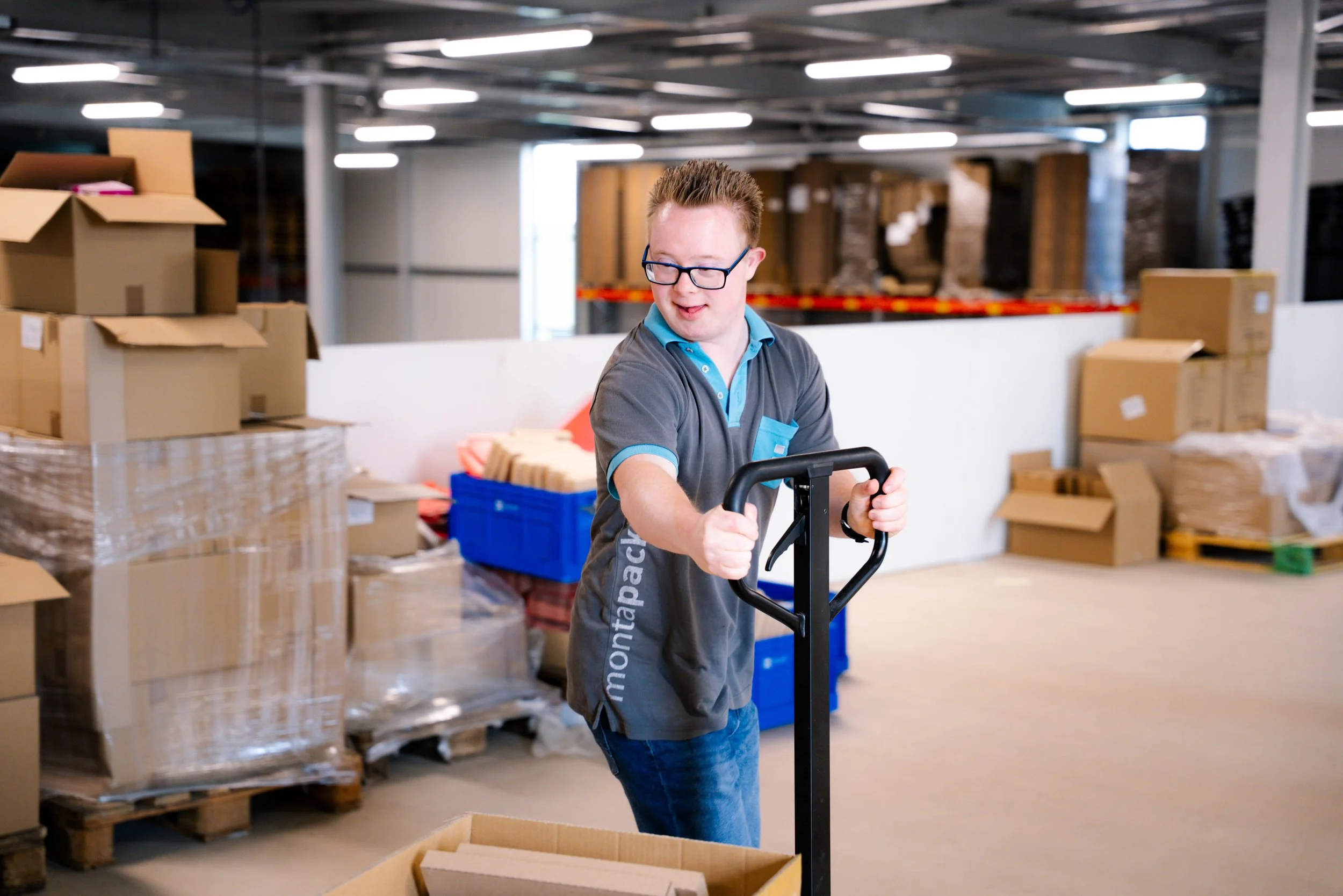 Een jonge man met een bril die een palletwagen duwt in een opslagruimte met dozen en verpakkingsmaterialen.