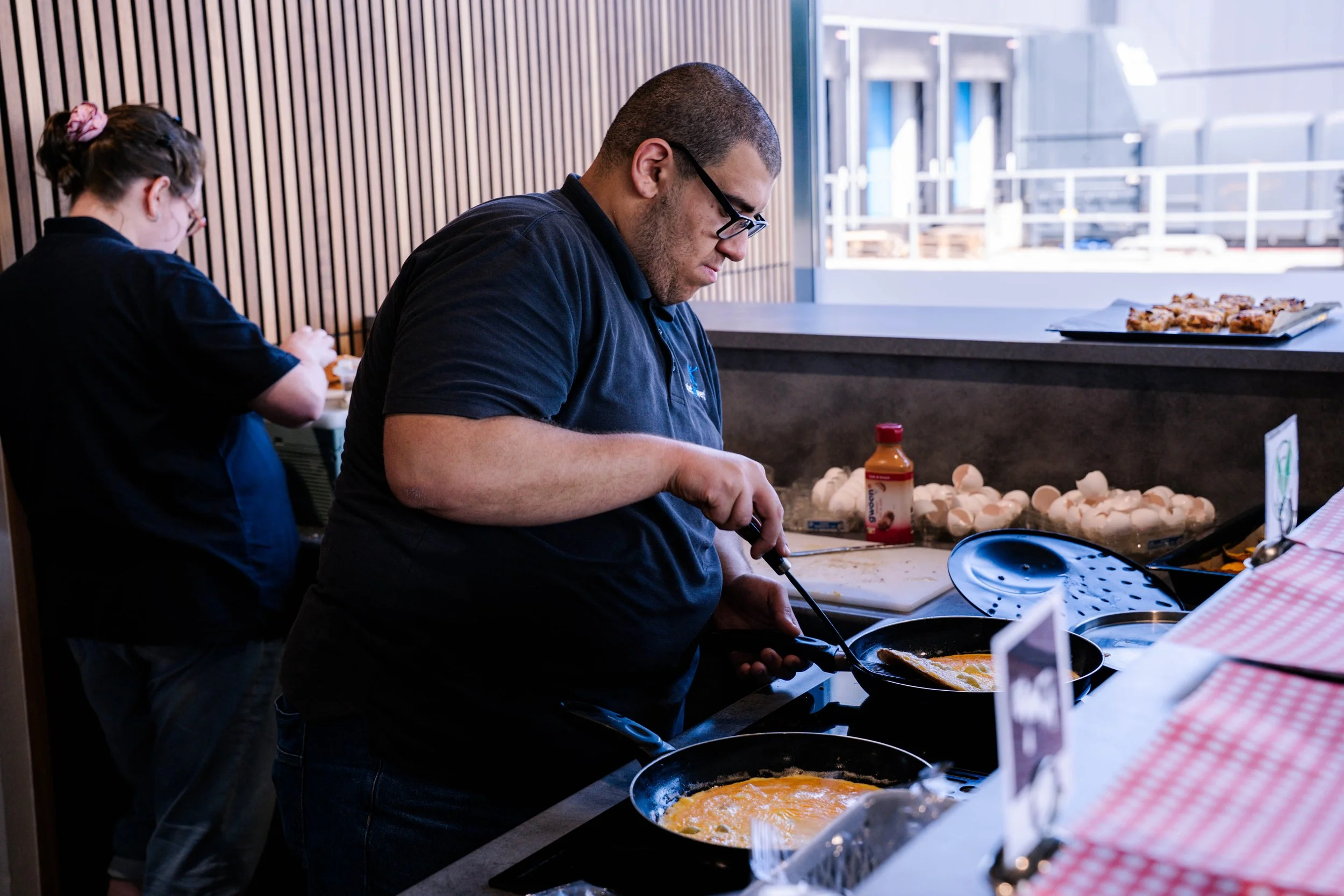 Man bereidt eten in een keuken, met een pan op het fornuis en een grote plaat met gebakken eieren naast zich.