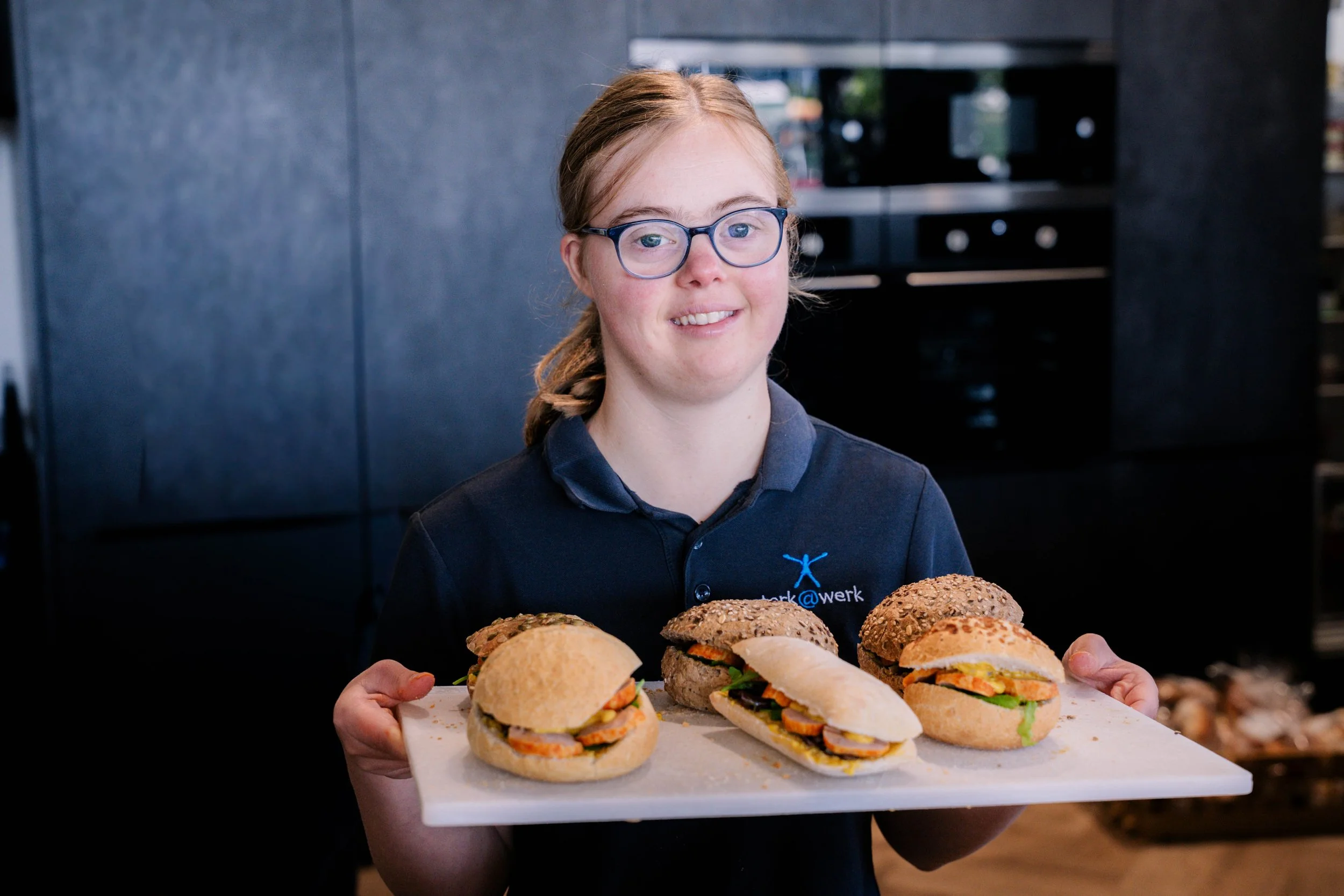 Vrouw met glazen en een donkerblauw uniform die een dienblad met verschillende belegde broodjes vasthoudt in een keuken.