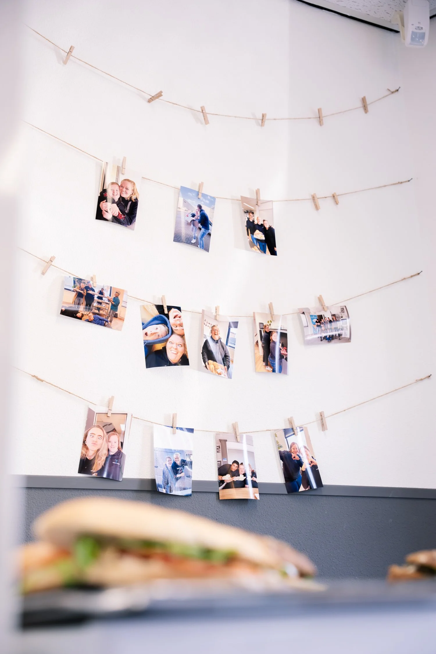 Foto van een muur met foto’s aan touwtjes vastgemaakt met houten knijpers. In de voorgrond zien we op de rand van een tafel een broodje met sla en vlees.