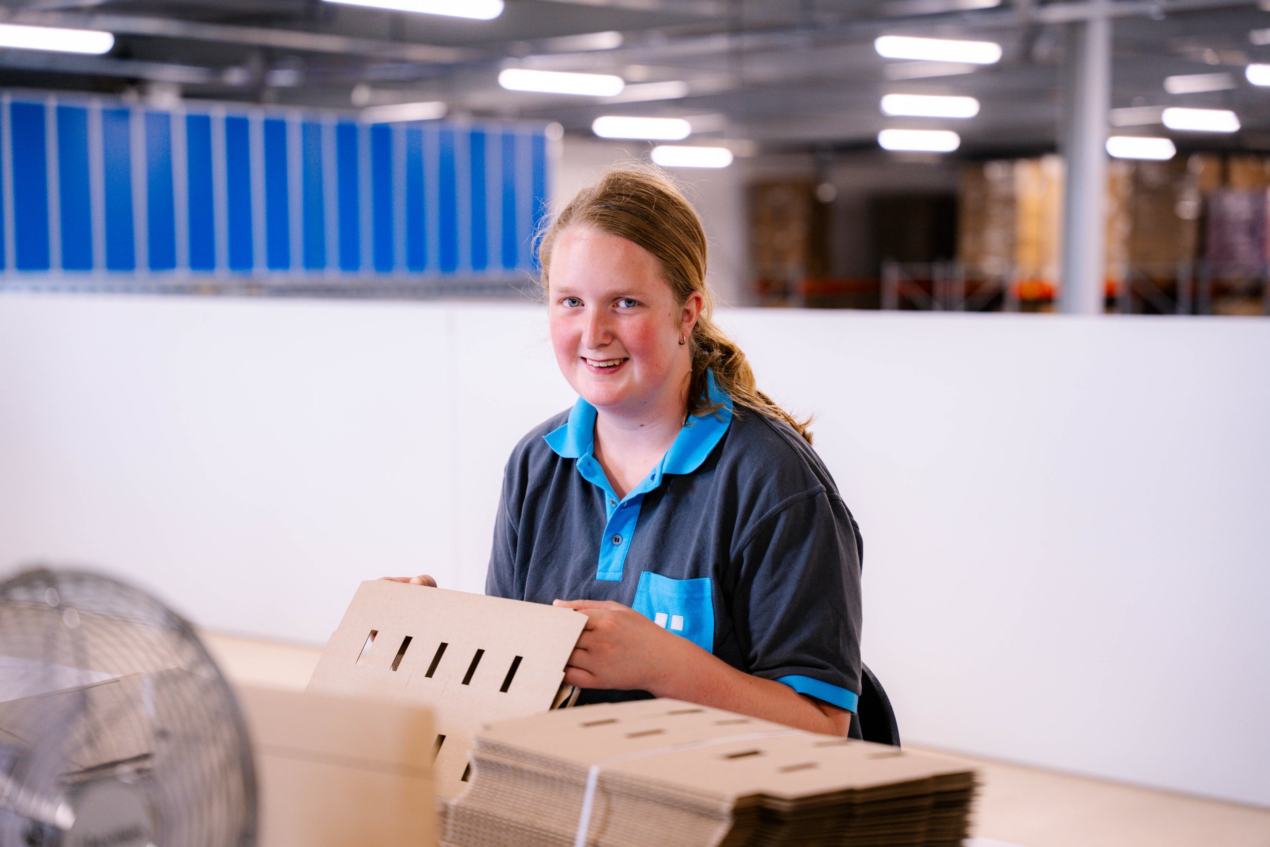Vrouw met rood haar en blauwe uniform die in een fabriek of magazijn een houten voorwerp vasthoudt, terwijl er in de buurt meerdere houten voorwerpen op een tafel liggen.