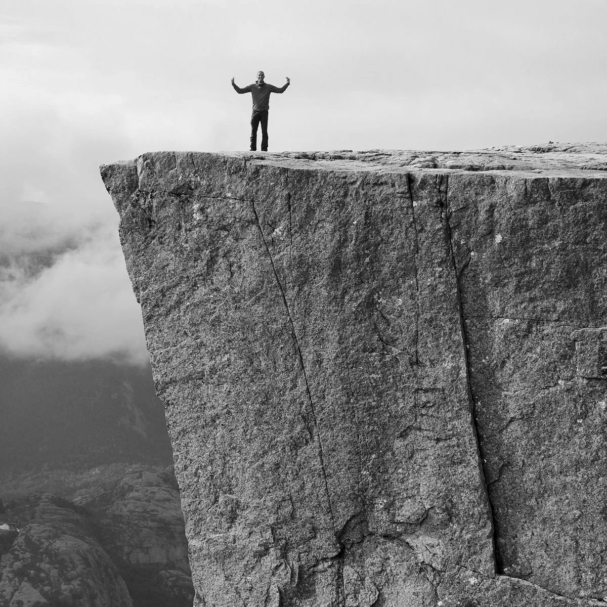 Een persoon staat op de rand van een grote rotsformatie met uitgestrekte armen, tegen een achtergrond van bewolkte bergen, in zwart-wit. Noorwegen. Mental health, coaching, welzijn, natuur, kalmte, groei, innerlijke rust