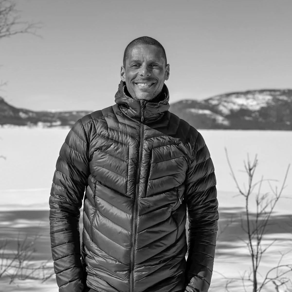 Man in een winterjas poseert buiten voor een bevroren meer en bergen op de achtergrond, op een snowy landschap, zwart-wit foto.