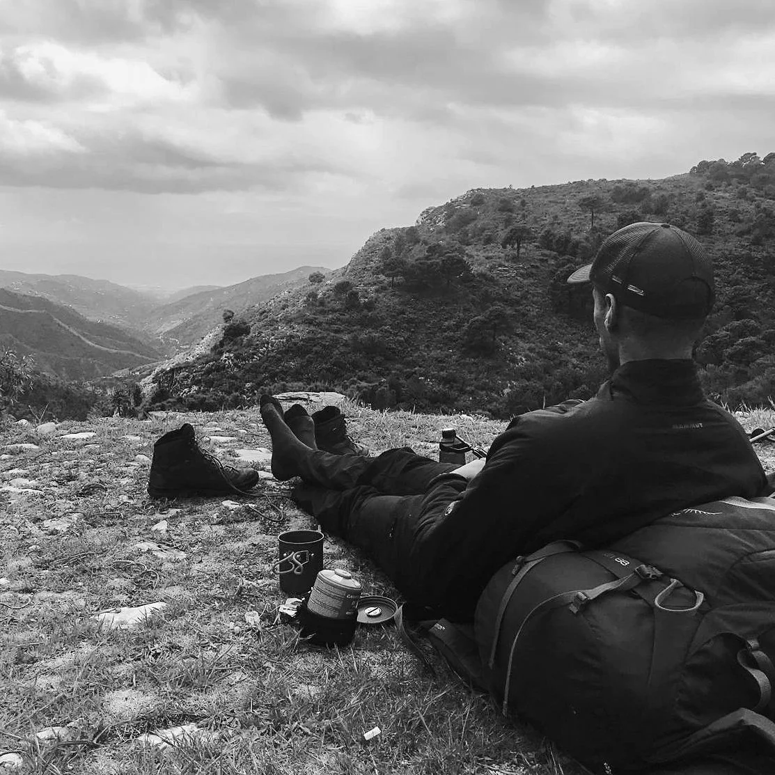 Man rust op een berg met uitzicht op mooie, glooiende heuvels en een bewolkte lucht. Mental health, coaching, welzijn, natuur, kalmte, groei, innerlijke rust